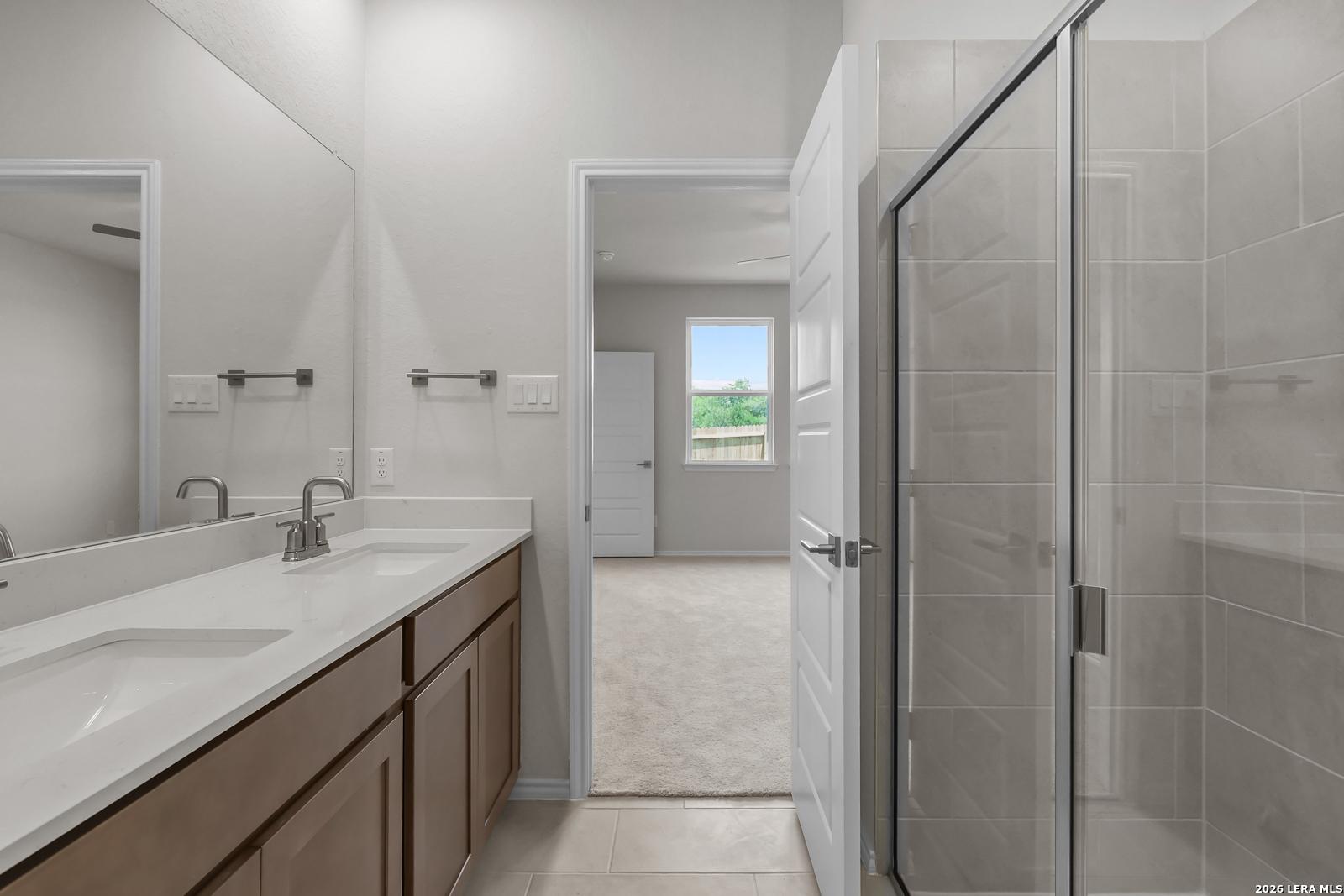 Modern bathroom with dual vanity, glass-enclosed shower, and bedroom view in The San Marcos F, Davidson Homes, San Antonio.
