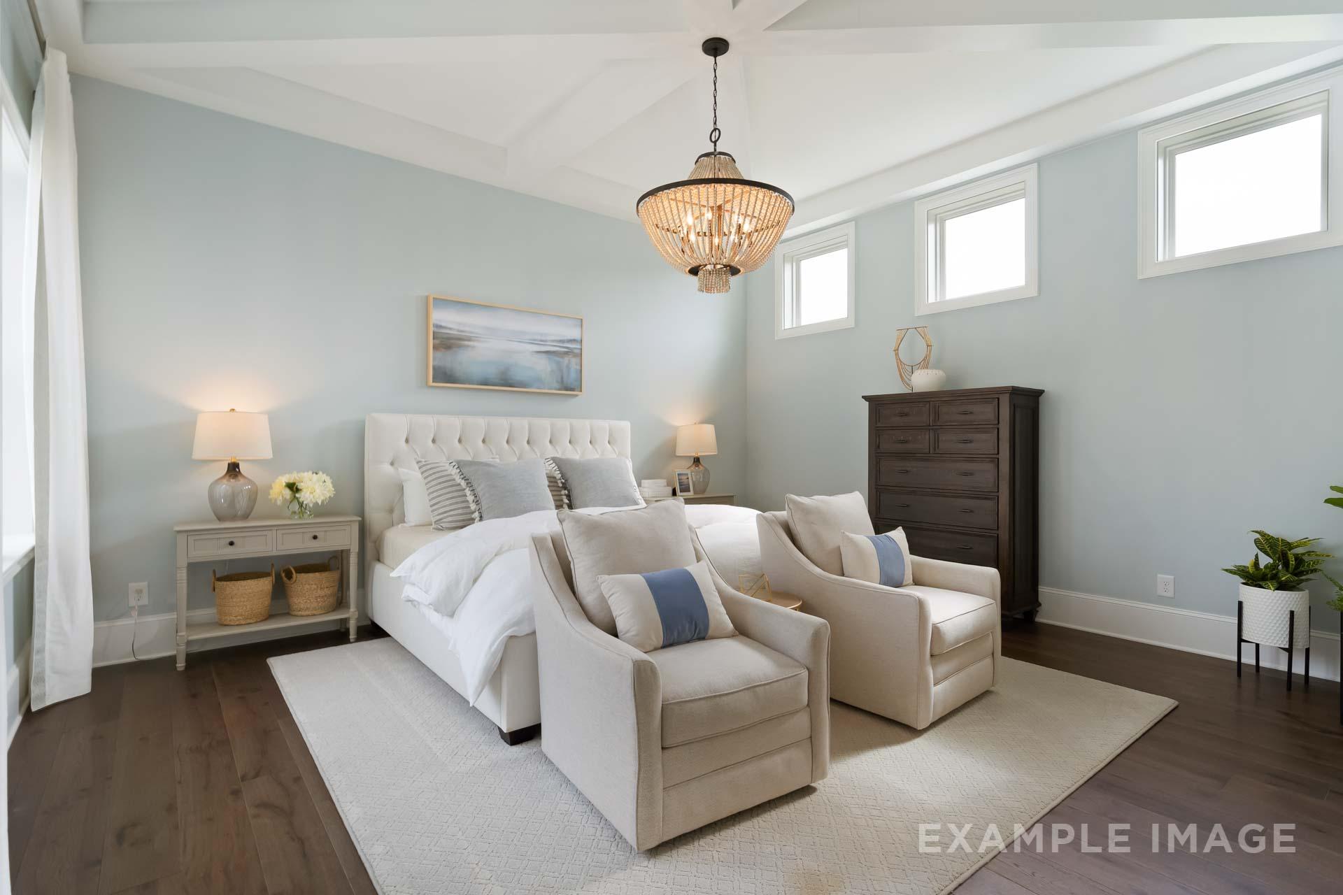 Upper floor master suite in The Seaside C with light blue walls, tufted white king bed, beige armchairs, wooden dresser, and beaded chandelier