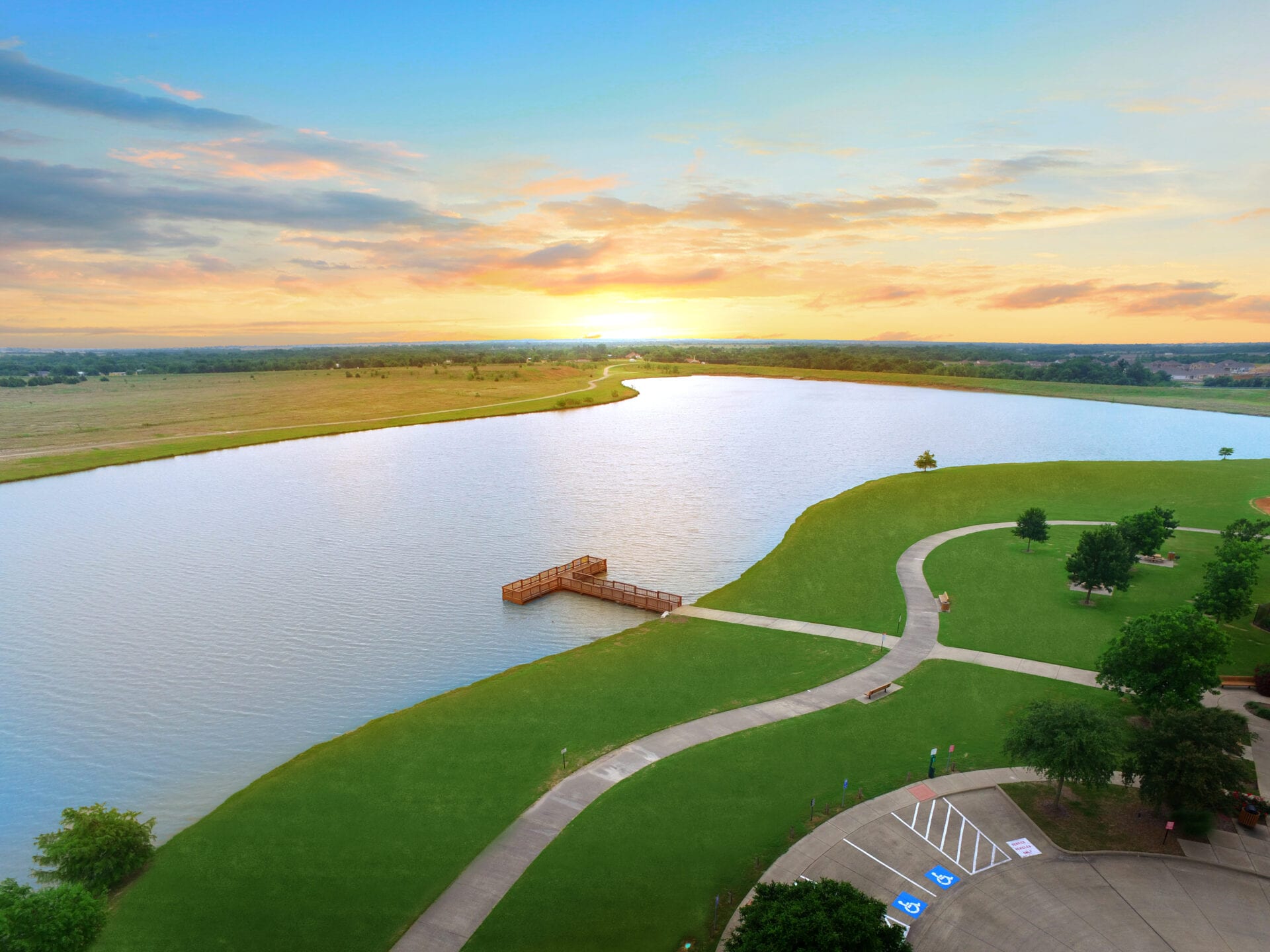 Aerial sunset view of serene lake with wooden dock, curved walking paths, green lawns and trees in Heartland Texas