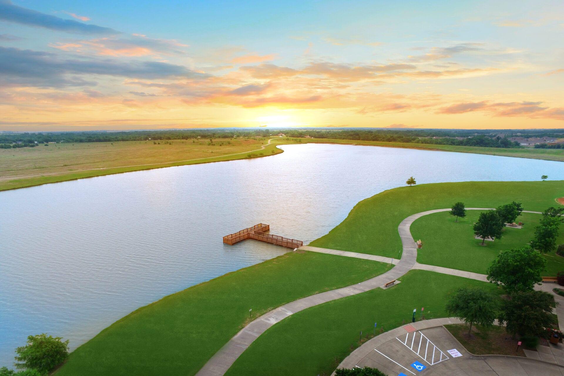 Aerial sunset view of serene lake with wooden dock, curved walking paths, green lawns and trees in Heartland Texas