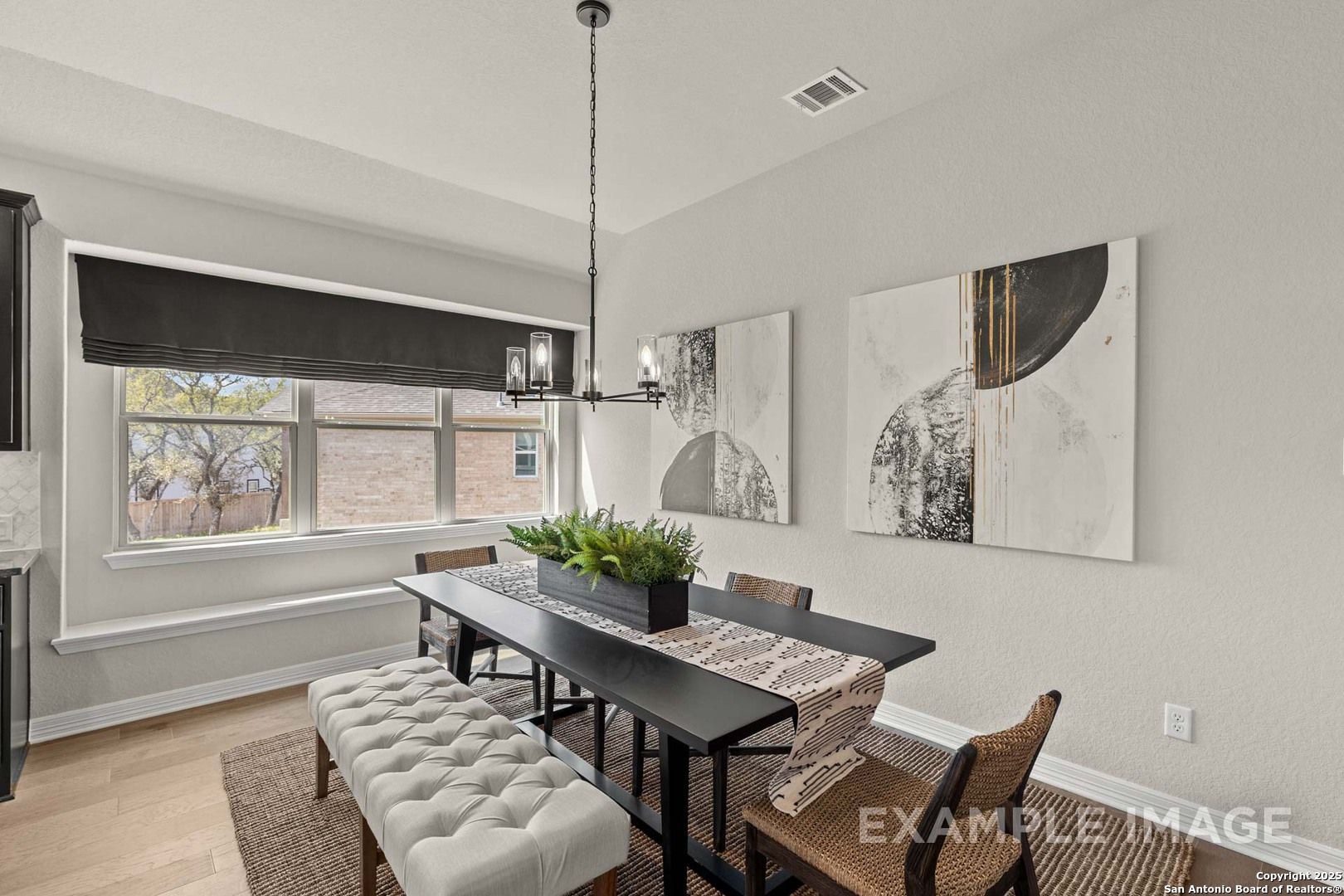 Modern dining room with large windows, chandelier, abstract art, wooden table, tufted bench, potted plant in The Summerlin B, Castroville, Texas