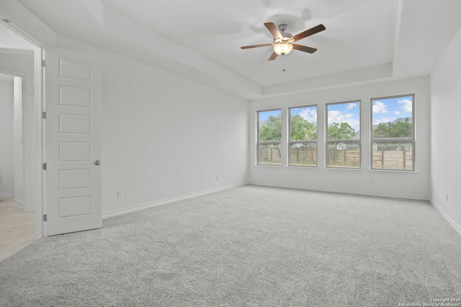 Bright living room with tray ceiling, ceiling fan, large windows, and gray carpet in Davidson Homes The Summerlin B, Castroville, Texas