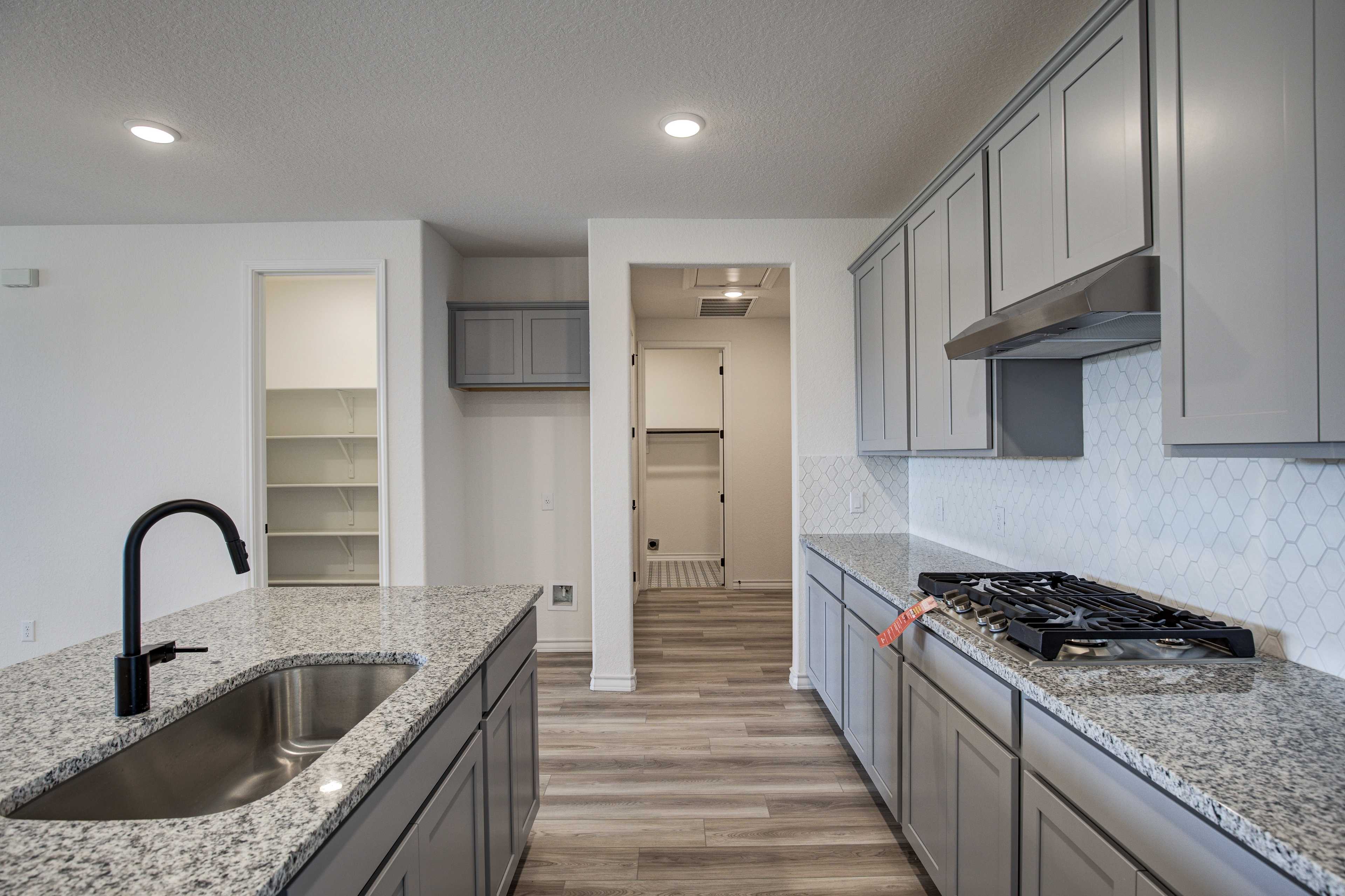 Spacious modern kitchen in The Lanier showcasing gray shaker cabinets, granite island with sink, gas range, and hexagonal tile backsplash