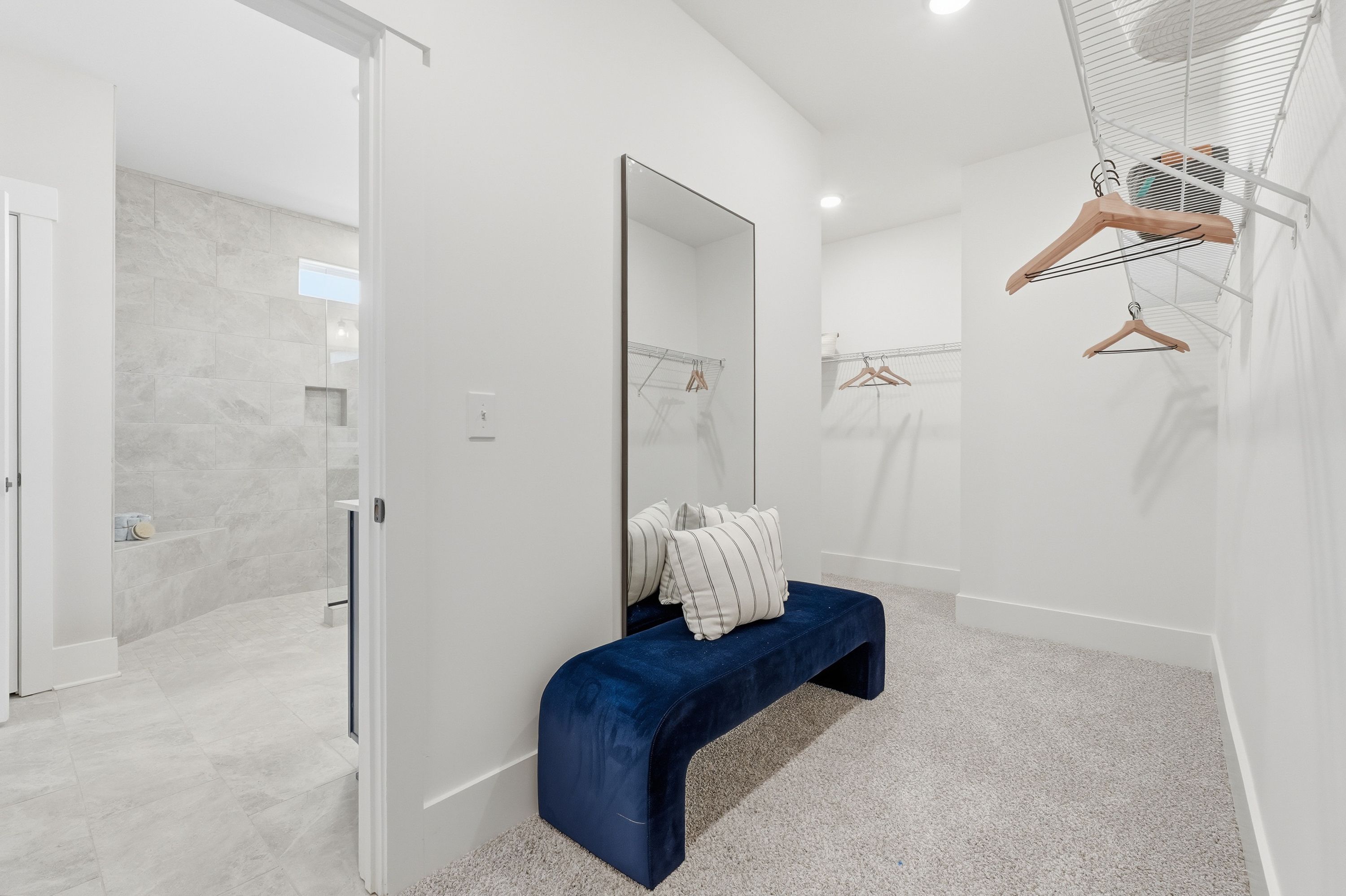Spacious walk-in closet at Noble Ridge in Cullman AL with white walls, hanging rods, full-length mirror, blue velvet bench, adjacent gray-tile bathroom