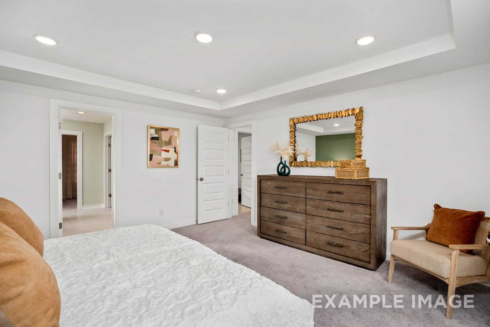 Spacious master bedroom in The Gordon B with white walls, wooden dresser, gold mirror, abstract art, plush white bed, and beige armchair