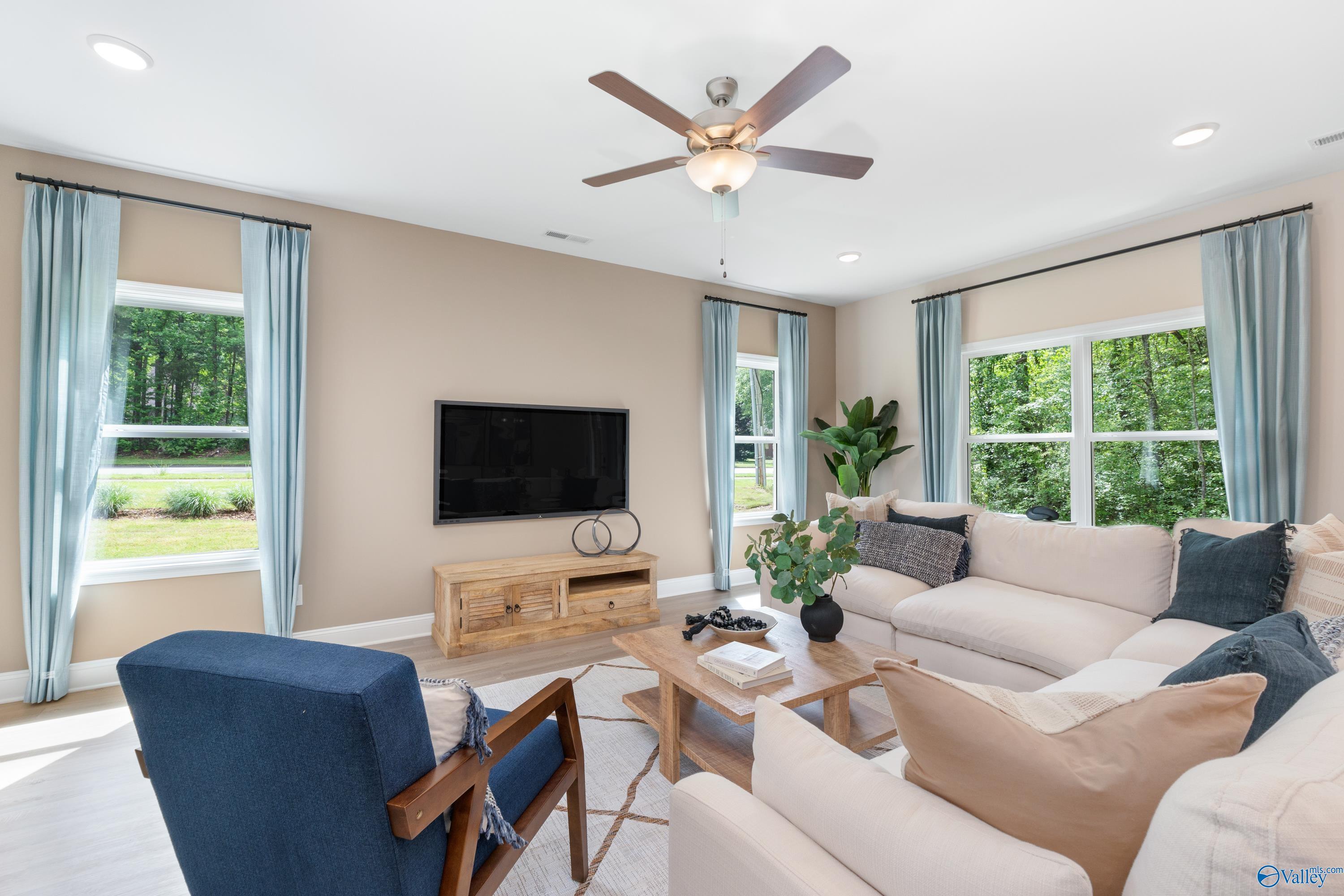Bright living room with beige sectional sofa, blue accent chair, wooden TV console, large windows overlooking greenery in Davidson Homes The Camden, Huntsville