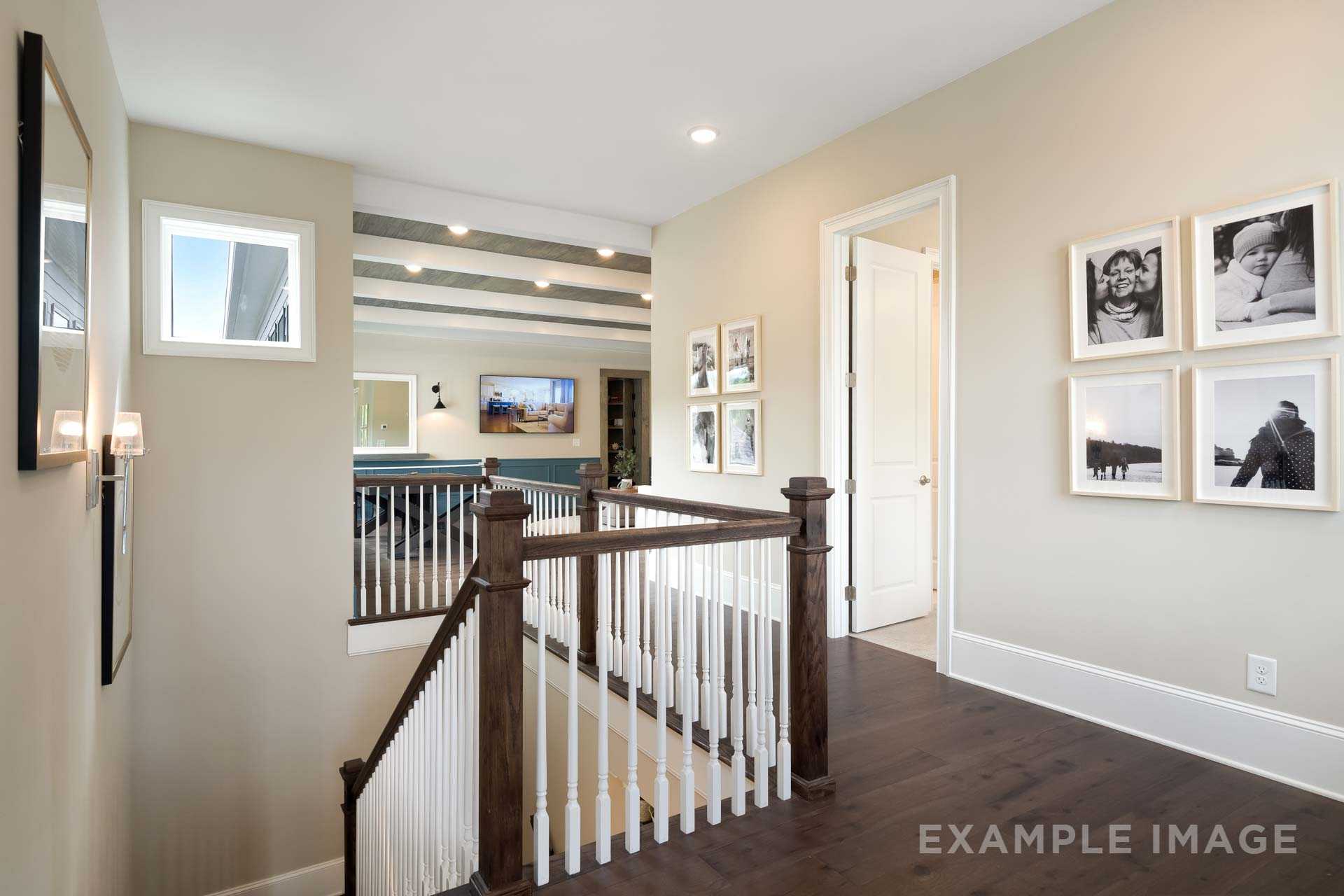 Upper floor hallway in The Seaside home design with hardwood floors, white baluster staircase, and framed photos on beige walls