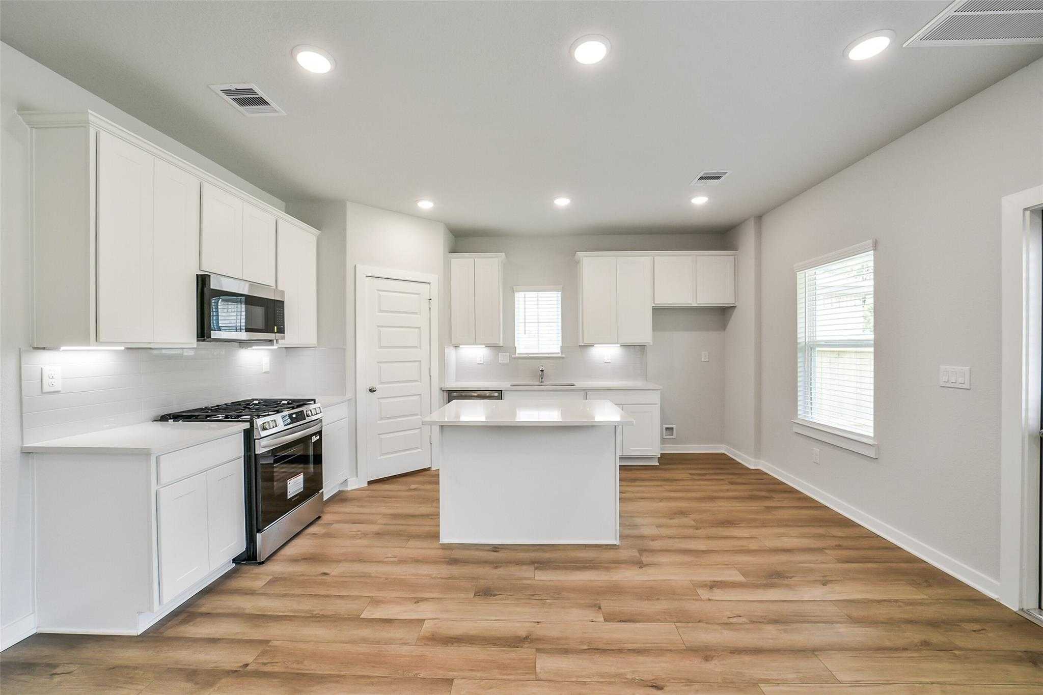 Modern white kitchen with island, stainless appliances, and hardwood floors in Davidson Homes The Trinity F, Magnolia, Texas
