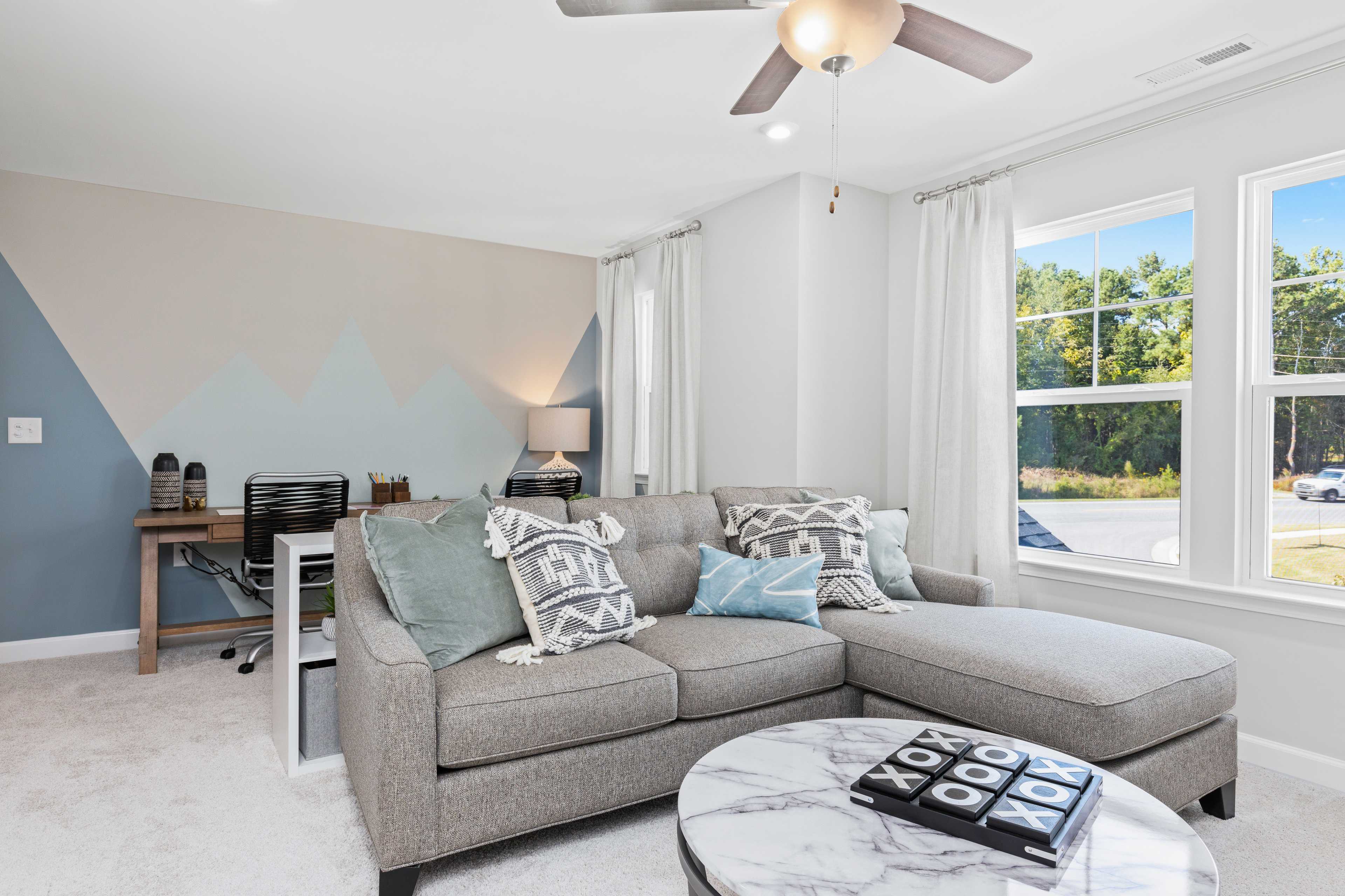 Cozy living room at Enclave at Belmont in Belmont NC with gray L-shaped sectional sofa, geometric accent wall, ceiling fan, corner desk, and large windows