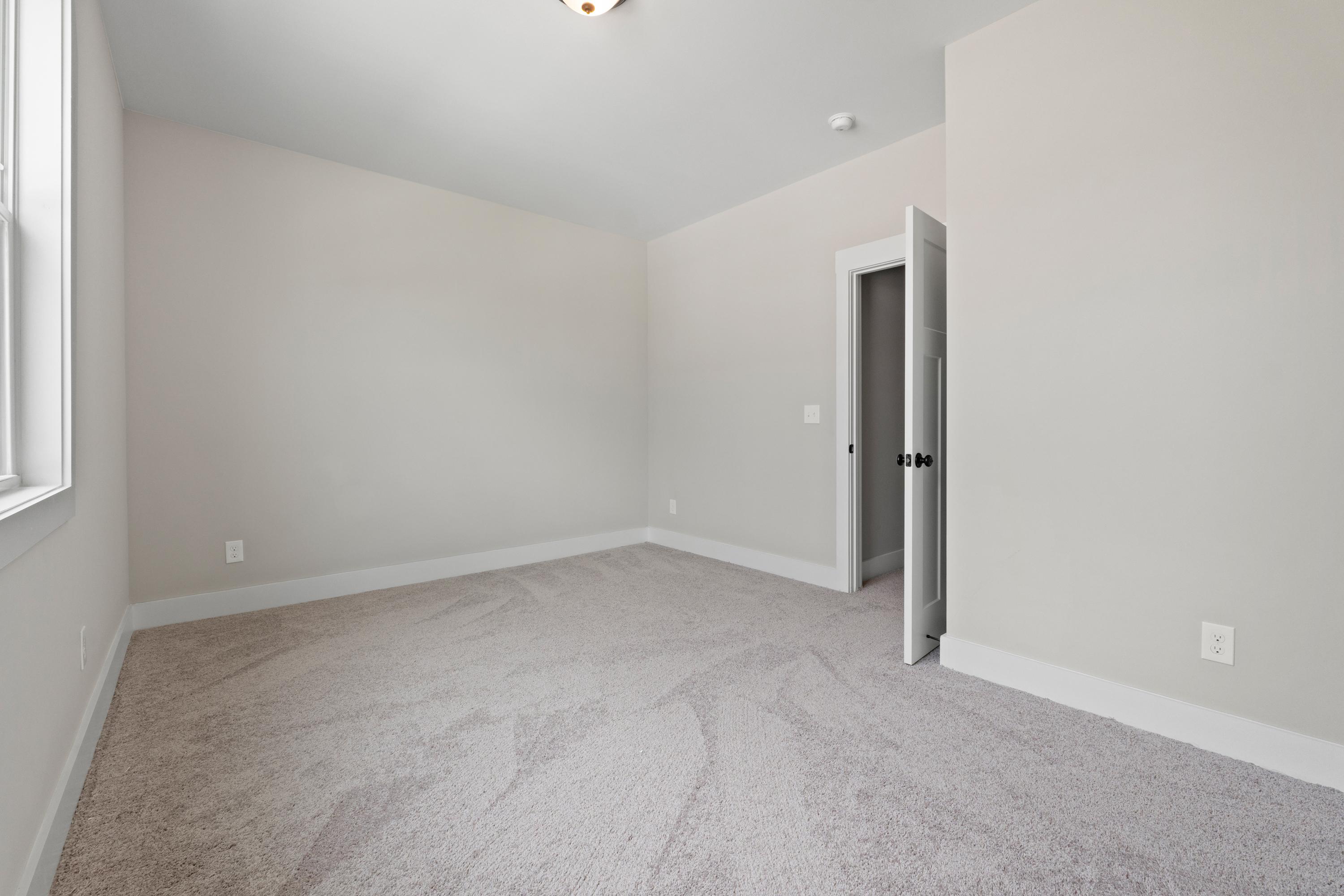 Spacious empty bedroom in The Oxford by Evermore Homes with neutral beige walls, gray carpet, large window, and open closet door
