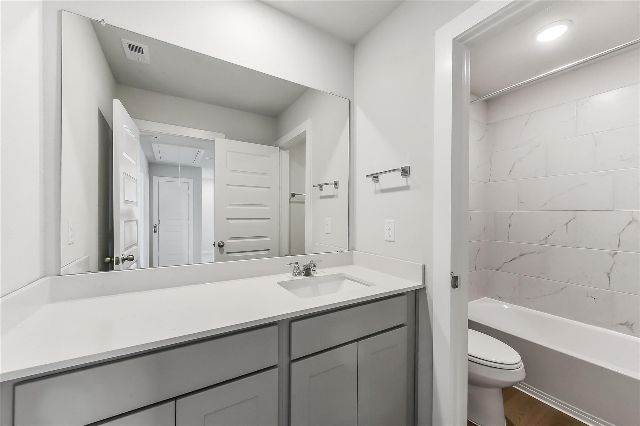 Modern guest bathroom featuring white subway tile tub surround, gray shaker vanity, and large mirror in 5-bedroom Davidson Homes The Brazos E, Magnolia, Texas