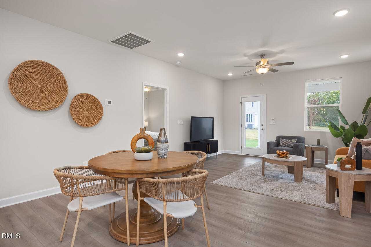 Bright open-concept living-dining area with round wood table, rattan chairs, woven wall decor, TV console, and sliding glass door in Davidson Homes Carter C, Lillington, NC