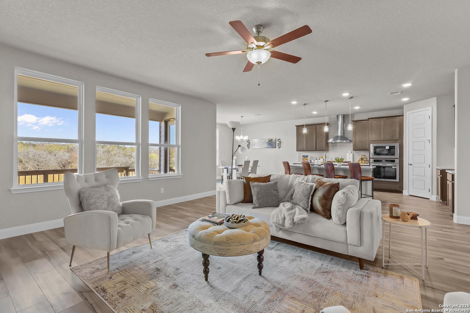 Cozy open-concept living room with beige sofa, armchair, ceiling fan, and kitchen view in Davidson Homes The Jennings G, Castroville, Texas
