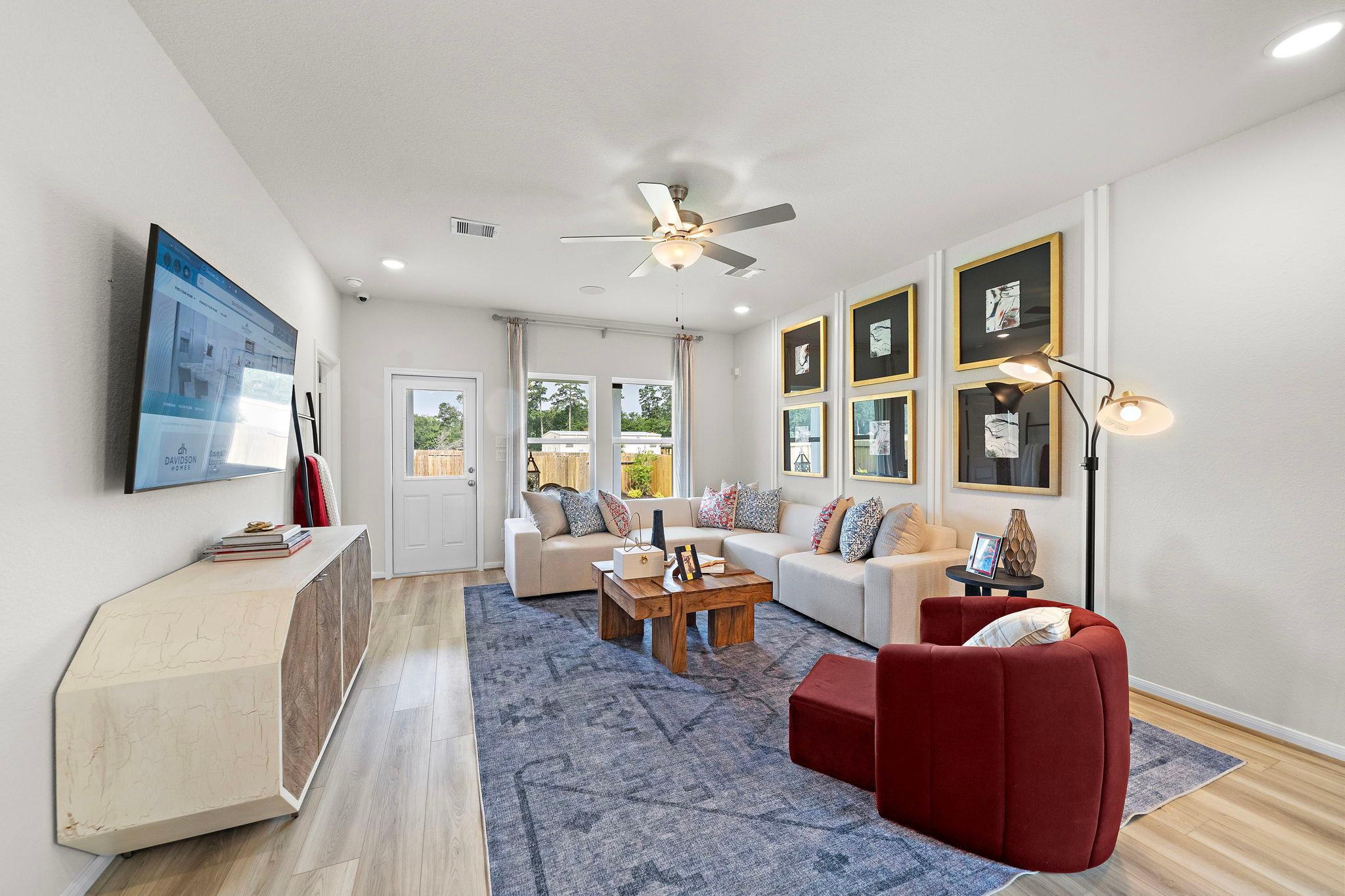 Spacious living room at Liberty Estates in Cleveland Texas with L-shaped sofa, hardwood floors, ceiling fan, wall art, and TV