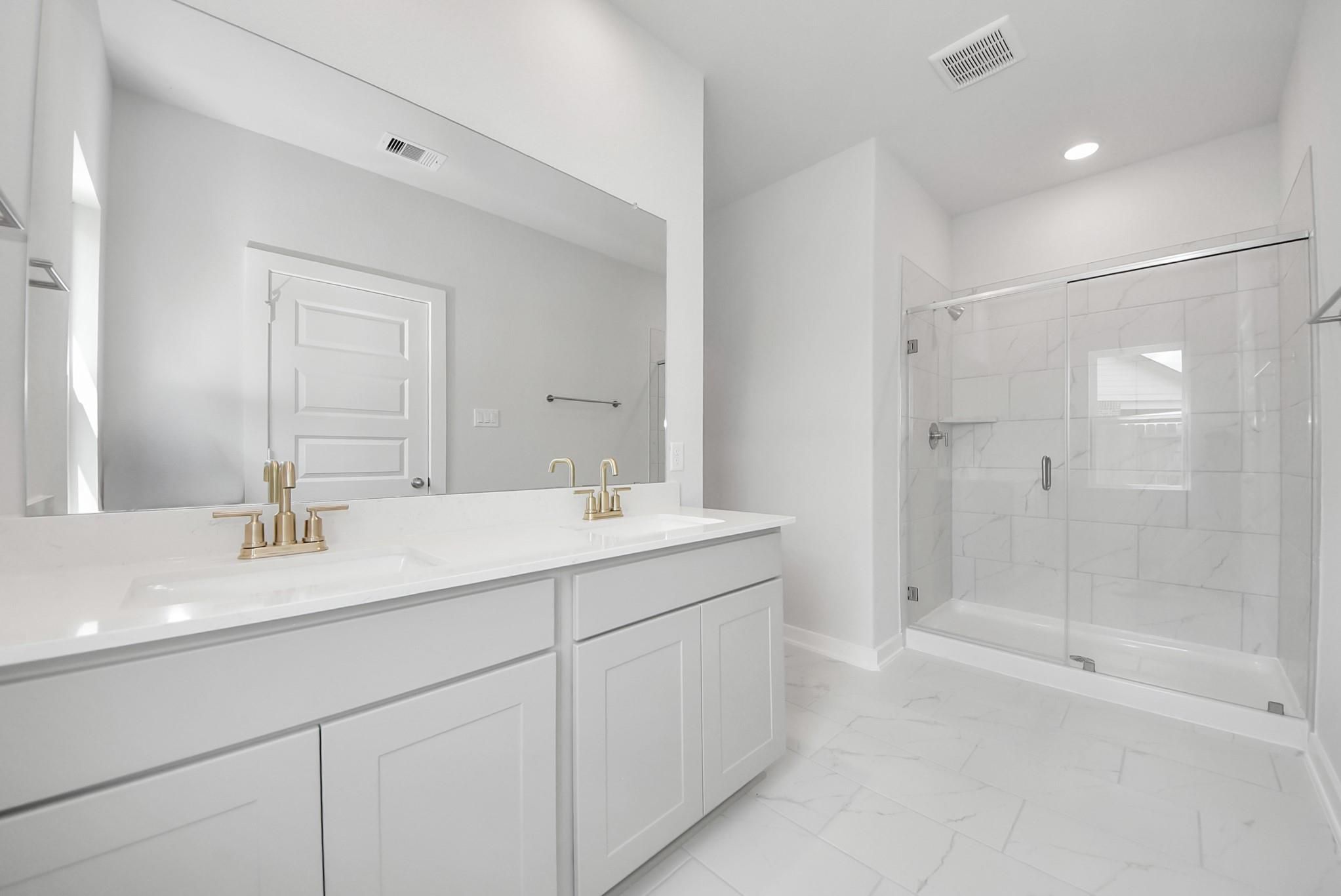 Modern primary bathroom with double vanity, gold faucets, and glass-enclosed walk-in shower in The Verano B, Sunterra, Katy.