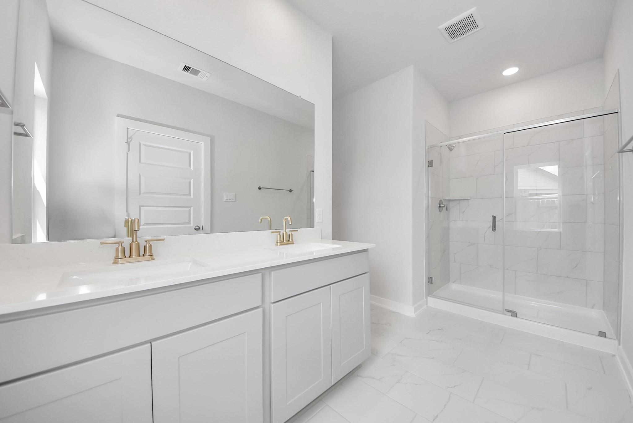 Modern primary bathroom with double vanity, gold faucets, and glass-enclosed walk-in shower in The Verano B, Sunterra, Katy.