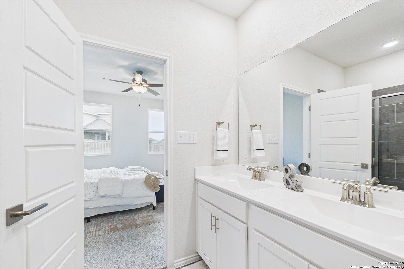 Modern master bathroom with double vanity, large mirror, and open bedroom view featuring ceiling fan in Davidson Homes The Asheville K, Bricewood, San Antonio