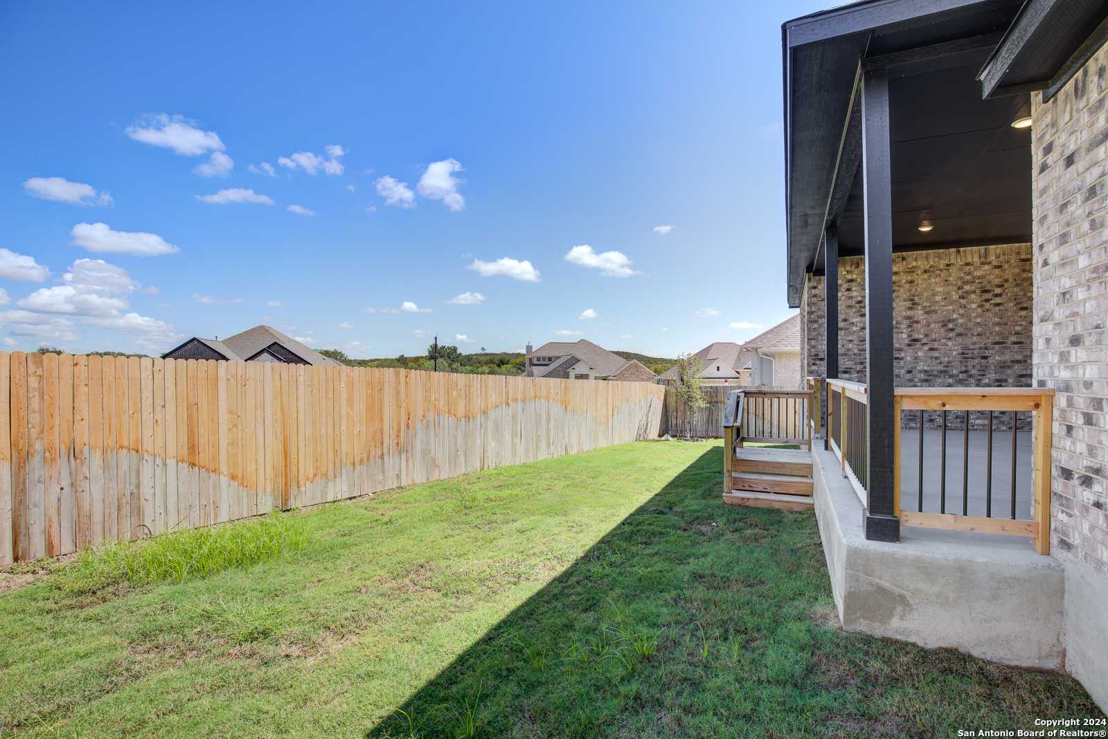 Covered back patio with wooden deck stairs, lush green lawn, and privacy fence in Davidson Homes The Garner B, Castroville, Texas
