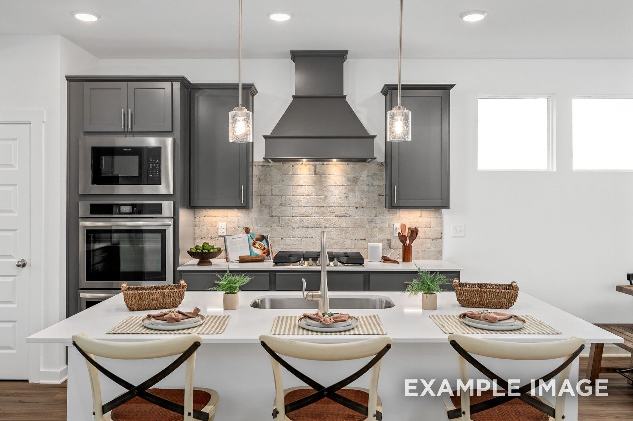 Modern kitchen with grey cabinets, brick backsplash, and large island in The Ash A floor plan at Woods Crossing, Gallatin.