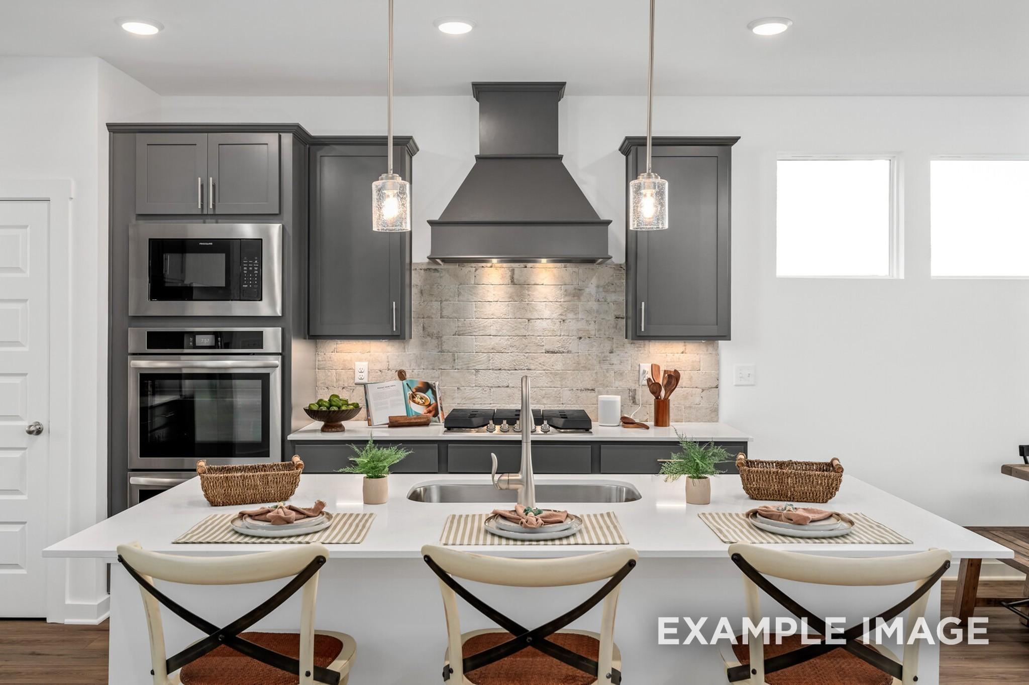Modern kitchen with grey cabinets, brick backsplash, and large island in The Ash A floor plan at Woods Crossing, Gallatin.