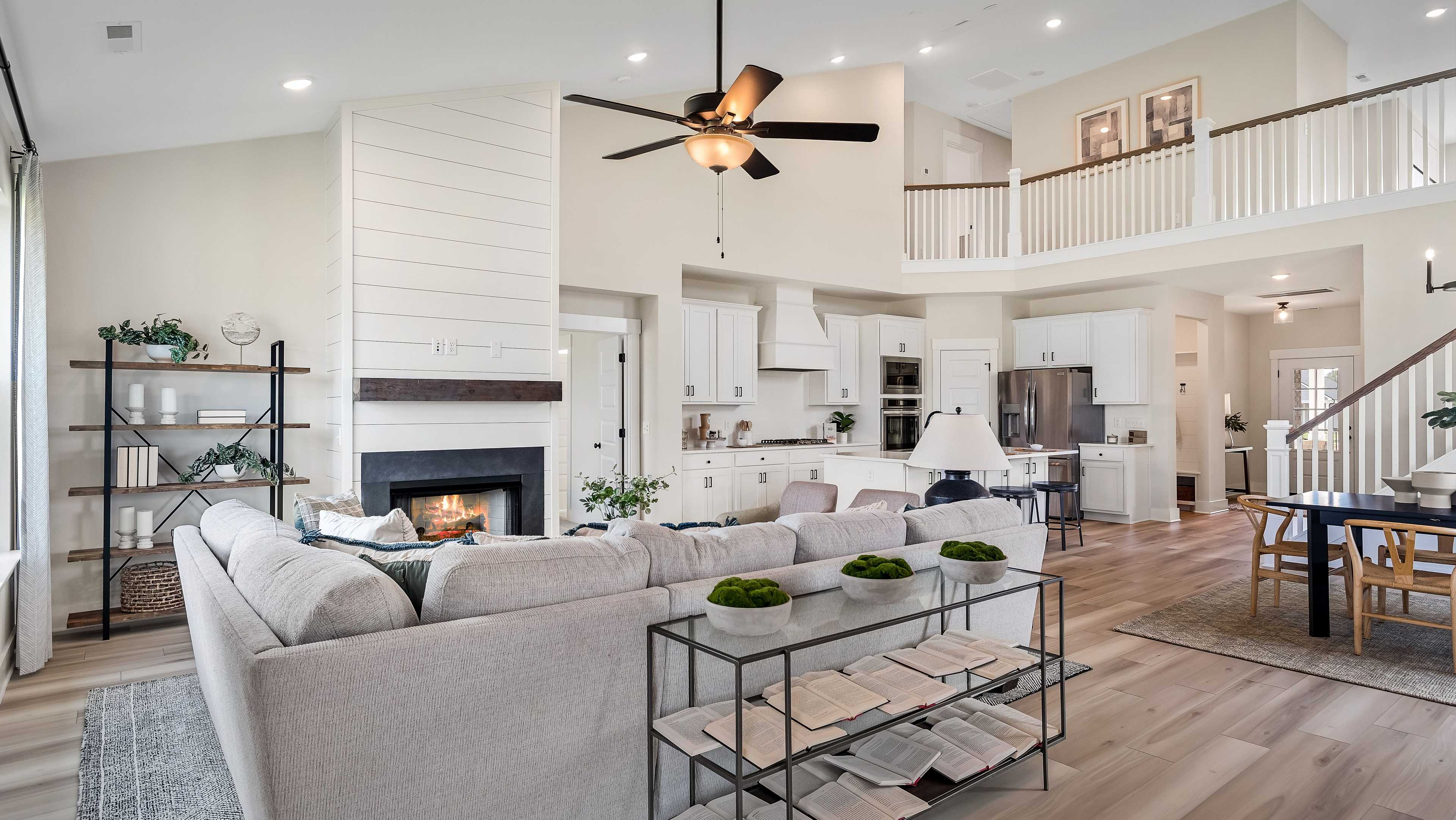Open floor plan with white shiplap fireplace, gray sectional sofa, modern kitchen cabinets, dining area, and loft balcony
