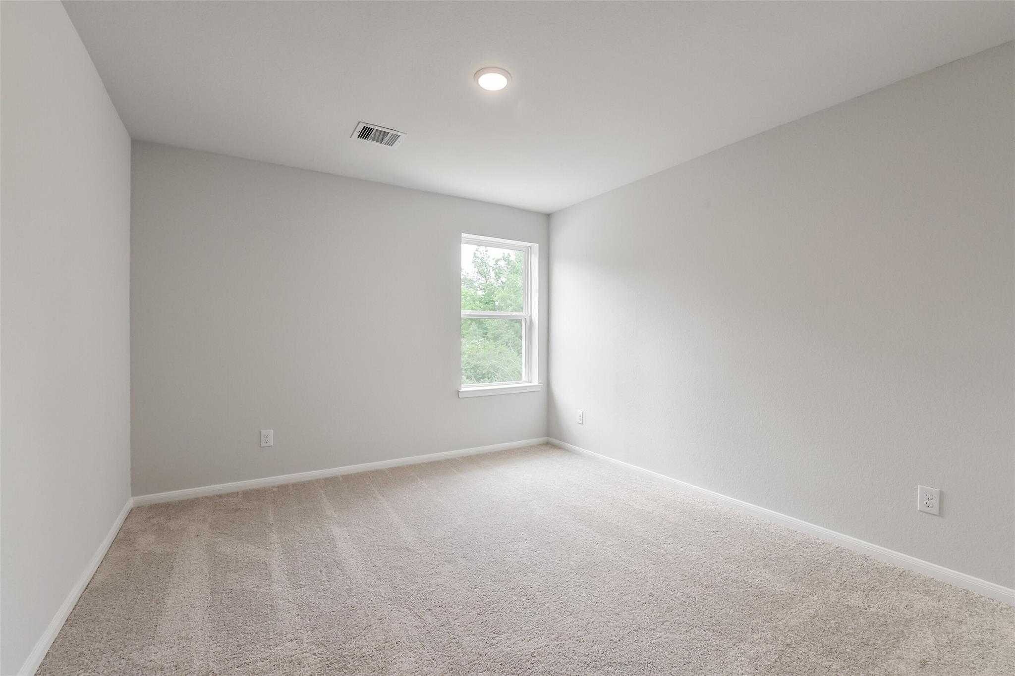 Spacious empty bedroom with light gray walls, beige carpet, ceiling light, and window in The Sabine E by Davidson Homes, Conroe, Texas