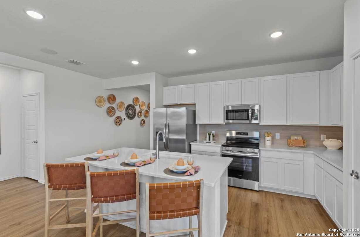 Modern white kitchen with stainless steel appliances, island bar stools, and woven wall decor in Davidson Homes The Daphne I, Bricewood, San Antonio