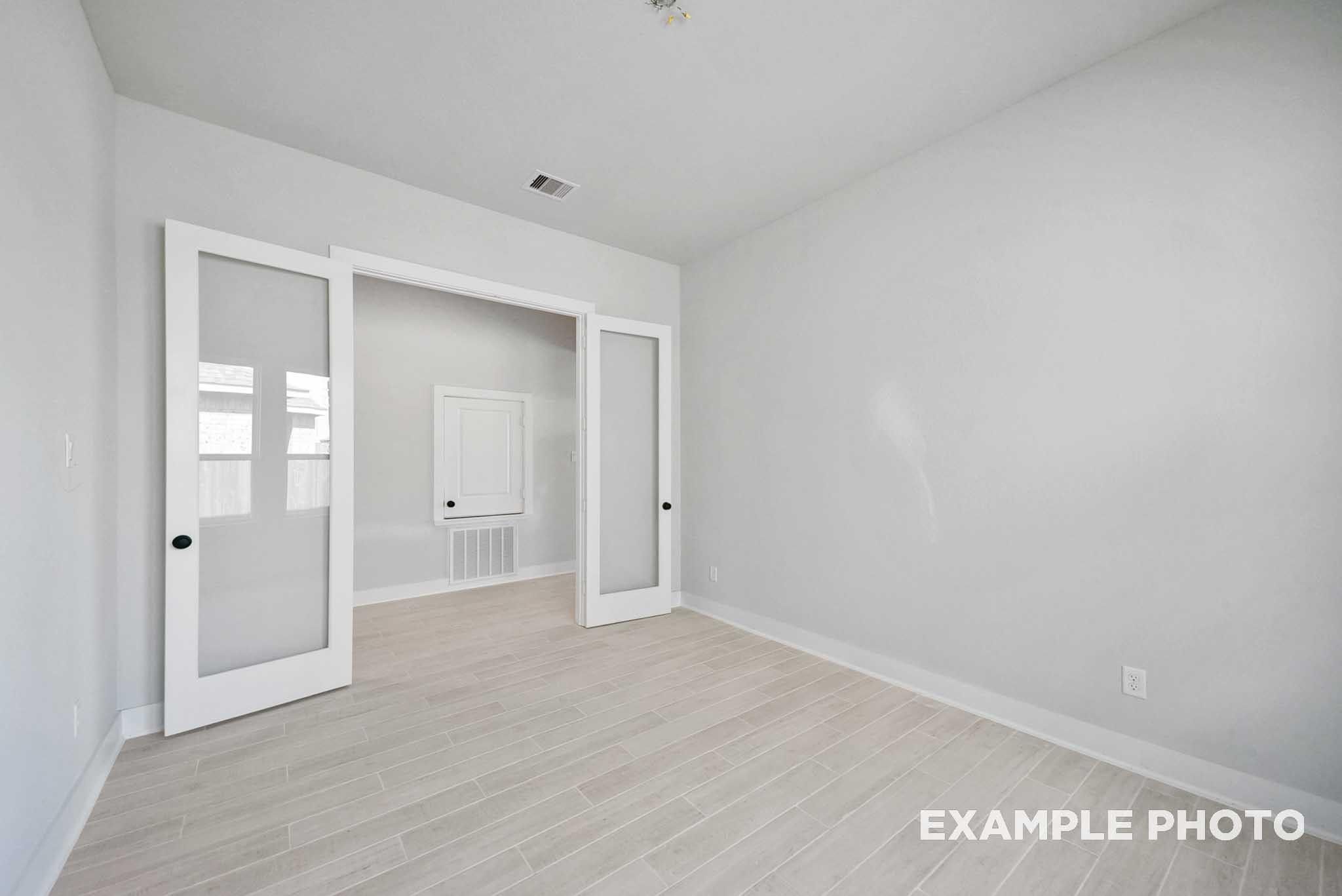 Bright bedroom with light oak flooring, white walls, and open glass French doors to closet in Davidson Homes The George A, Texas City