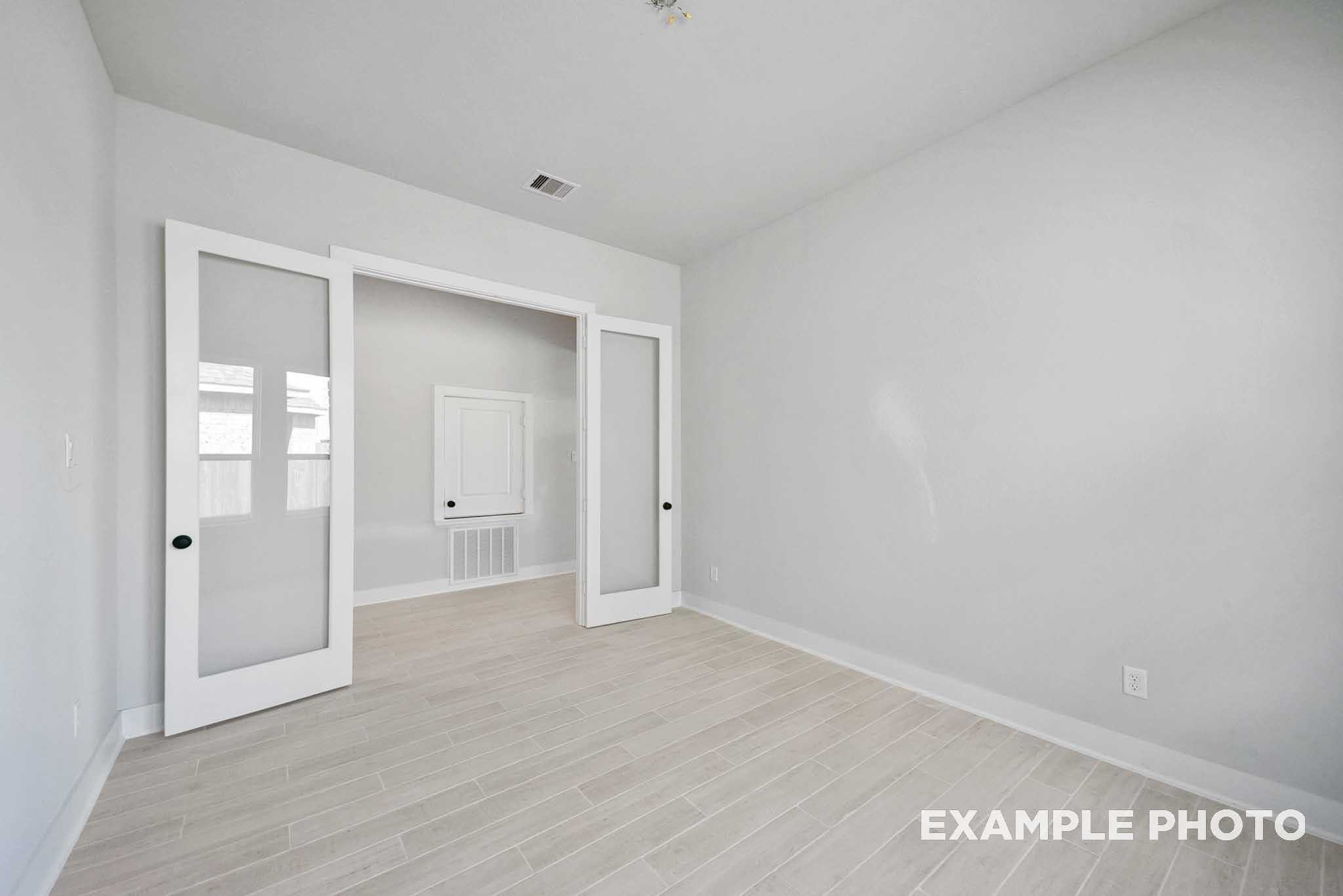 Bright bedroom with light oak flooring, white walls, and open glass French doors to closet in Davidson Homes The George A, Texas City