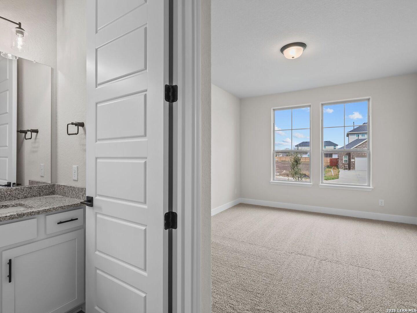 Bright bedroom with carpet and attached bathroom featuring granite vanity in The Belmont H floor plan, Castroville, TX.