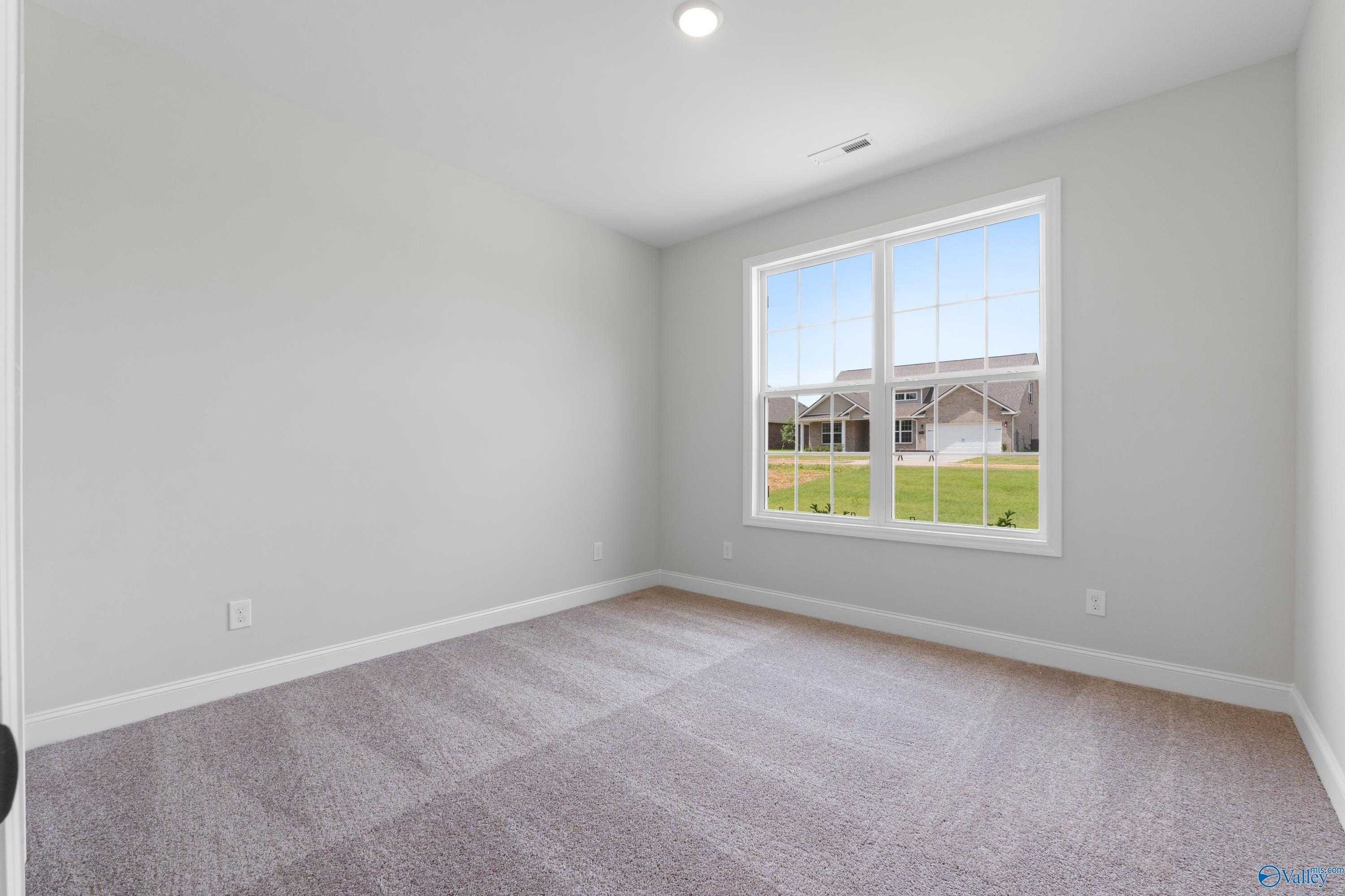 Empty bedroom with light gray walls, beige carpet floor, and large window overlooking green lawn in Davidson Homes The Kirkland, Harvest, Alabama