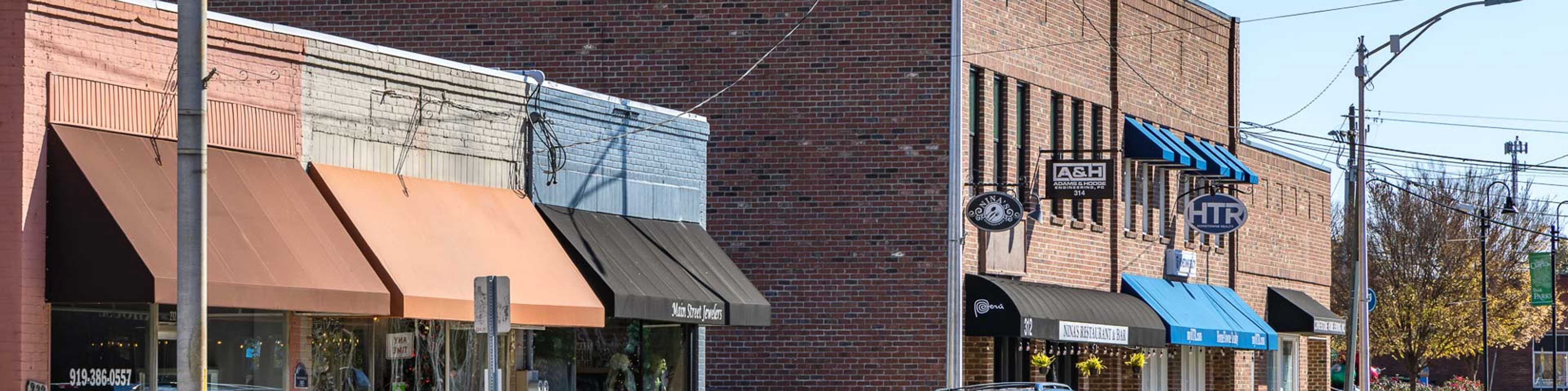Quaint brick storefronts with colorful awnings line sunny street in Clayton community, parked cars and fall trees
