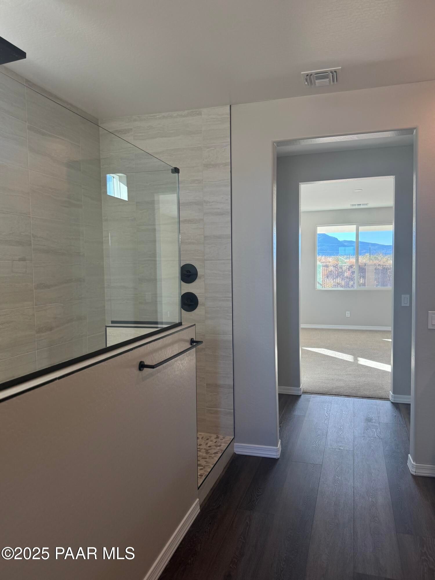 Modern master bathroom with frameless glass walk-in shower, pebble tile floor, and mountain view from adjacent bedroom in The Monarch E, Prescott Arizona