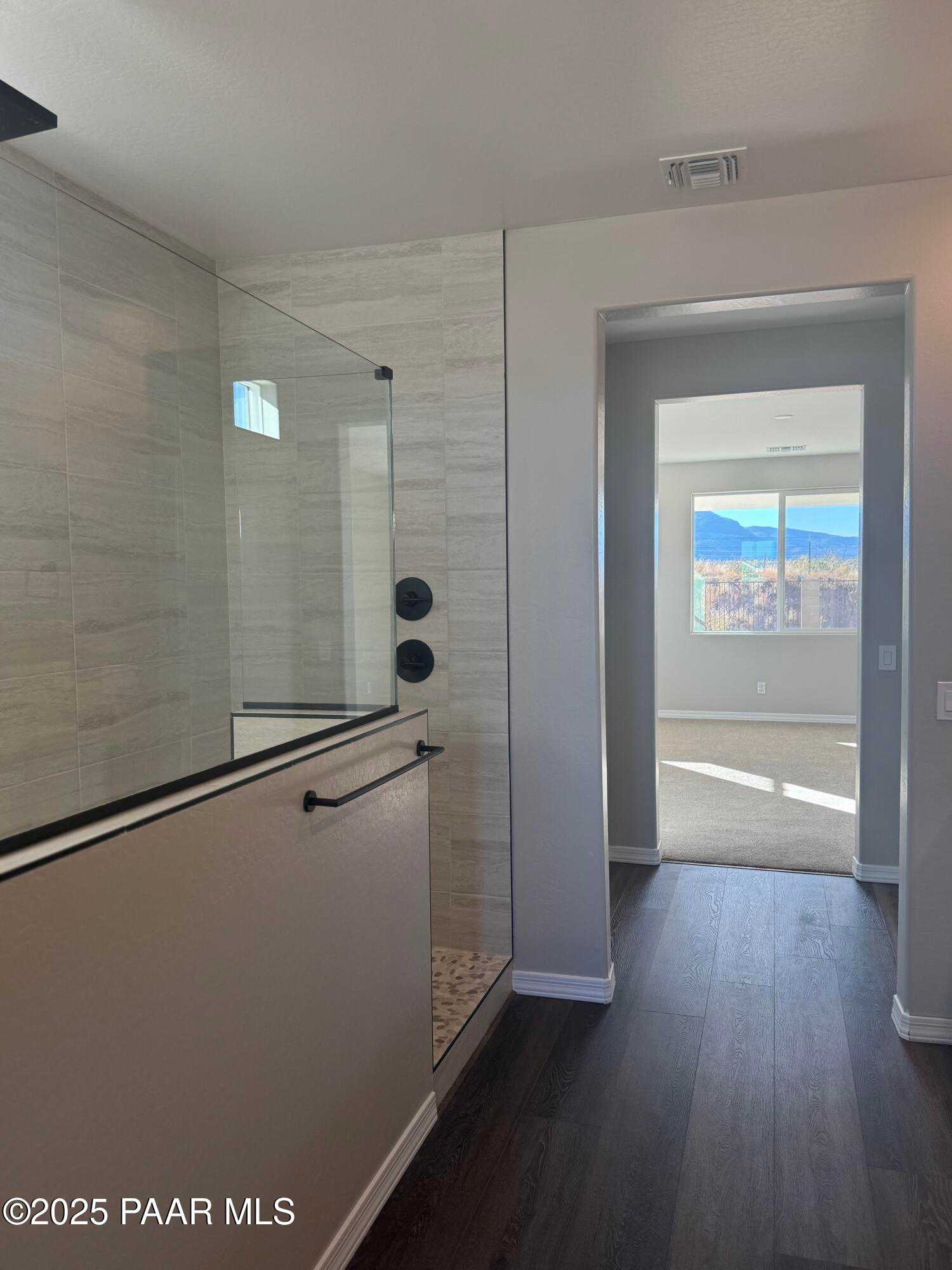 Modern master bathroom with frameless glass walk-in shower, pebble tile floor, and mountain view from adjacent bedroom in The Monarch E, Prescott Arizona