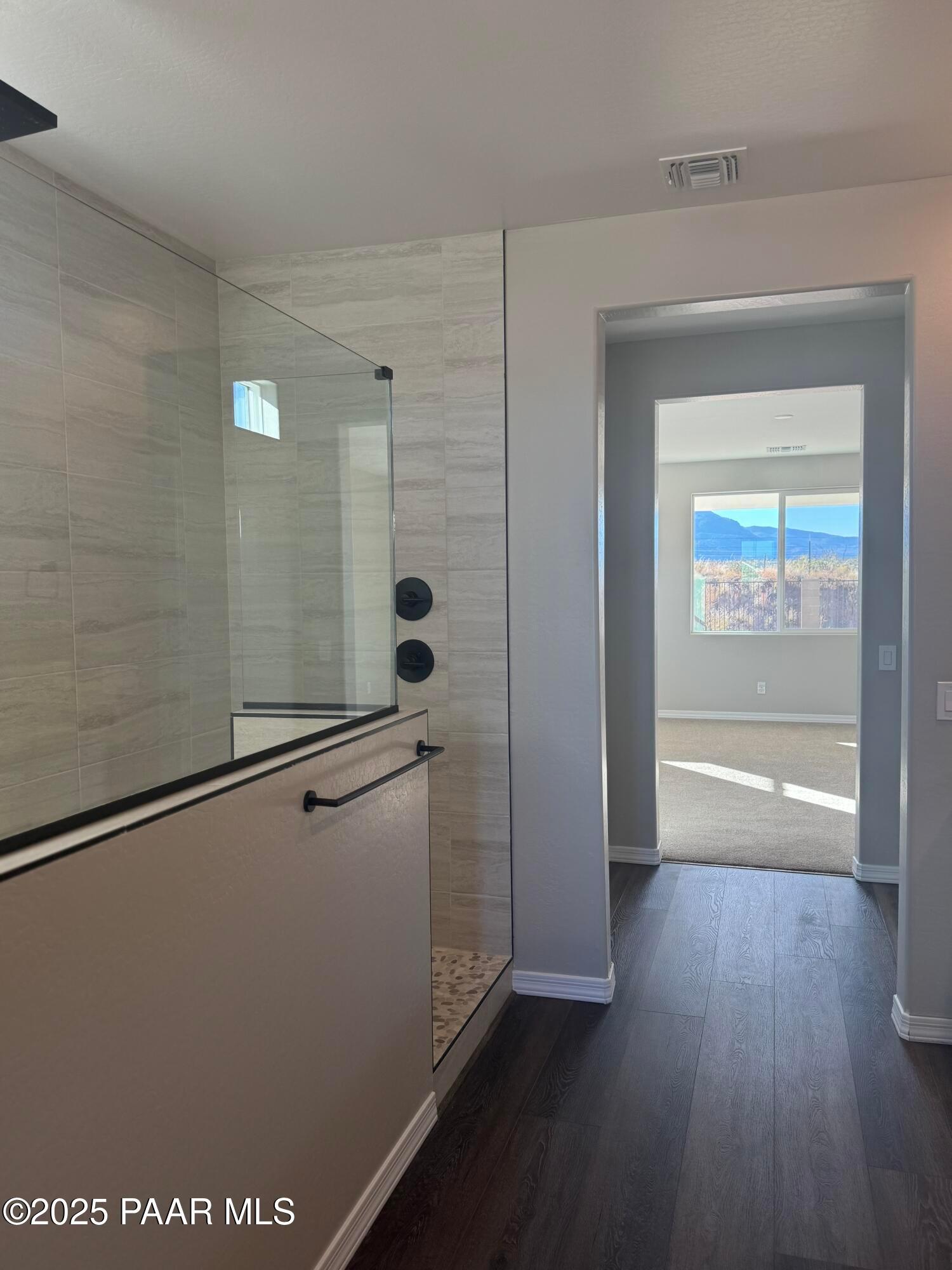 Modern master bathroom with frameless glass walk-in shower, pebble tile floor, and mountain view from adjacent bedroom in The Monarch E, Prescott Arizona