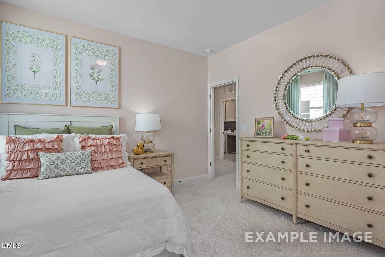 Cozy pink bedroom with white king bed, ruffled pillows, wooden dresser, round mirror, botanical art in 5-bed Davidson Homes Hickory II C, Wake Forest NC