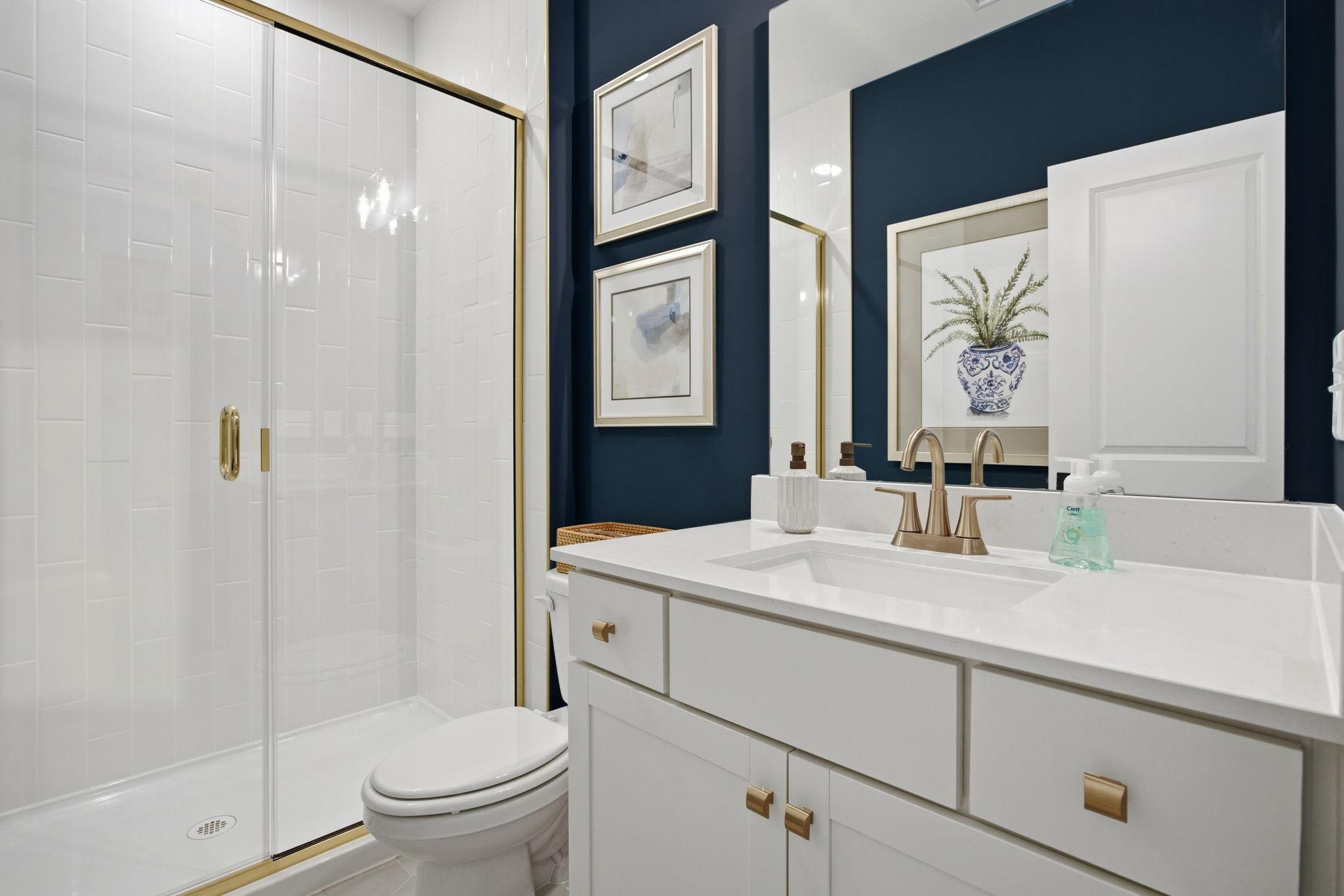 Modern bathroom in The Ashport K featuring a white vanity, gold fixtures, and a tiled walk-in shower against navy walls.