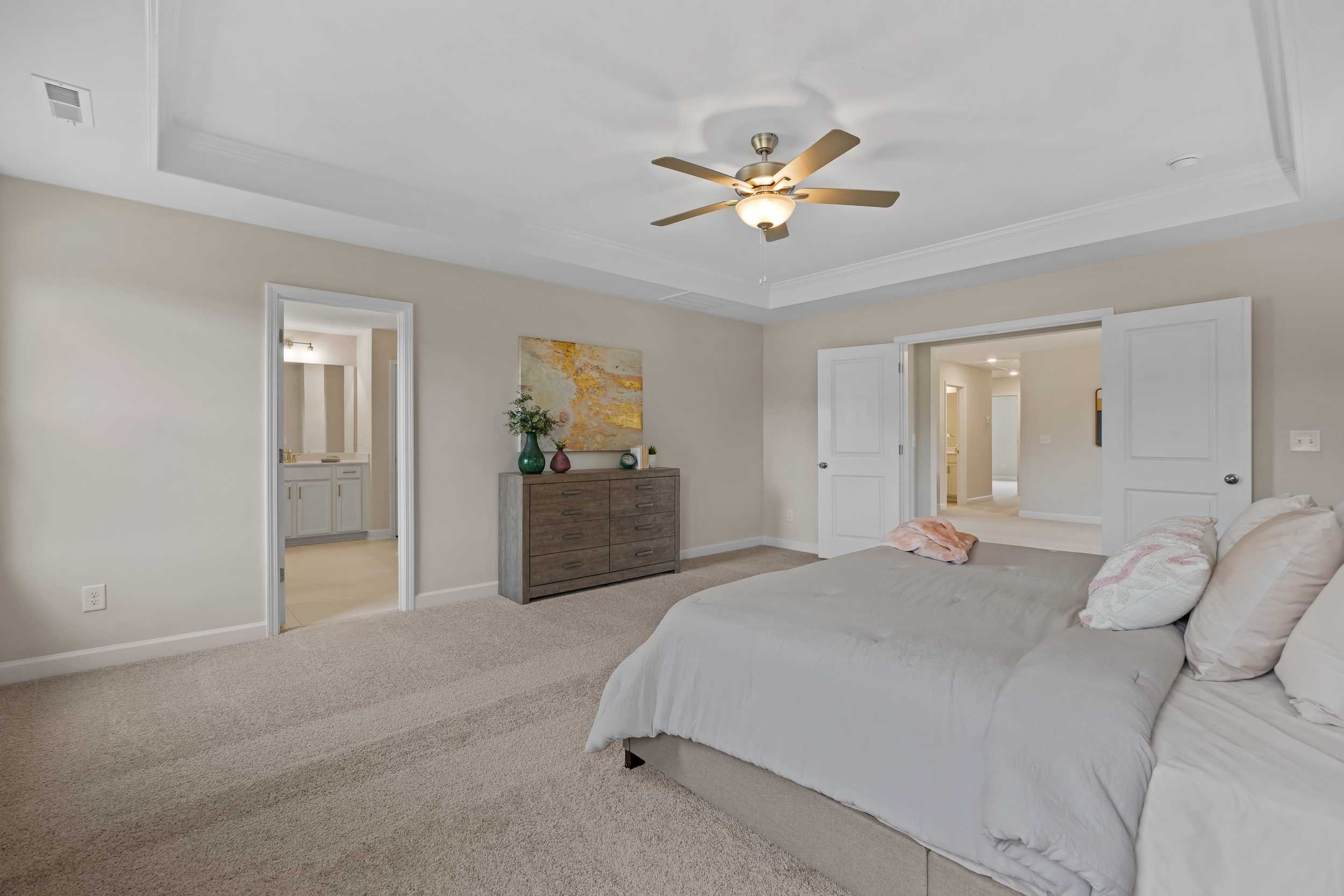 Spacious master bedroom in Gregory Village, Lillington NC by Davidson Homes with king bed, beige walls, ceiling fan, and en-suite bath