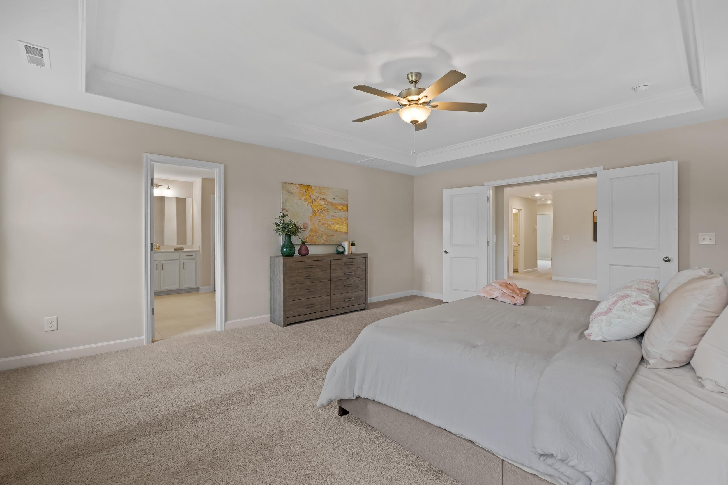 Spacious master bedroom in Gregory Village, Lillington NC by Davidson Homes with king bed, beige walls, ceiling fan, and en-suite bath