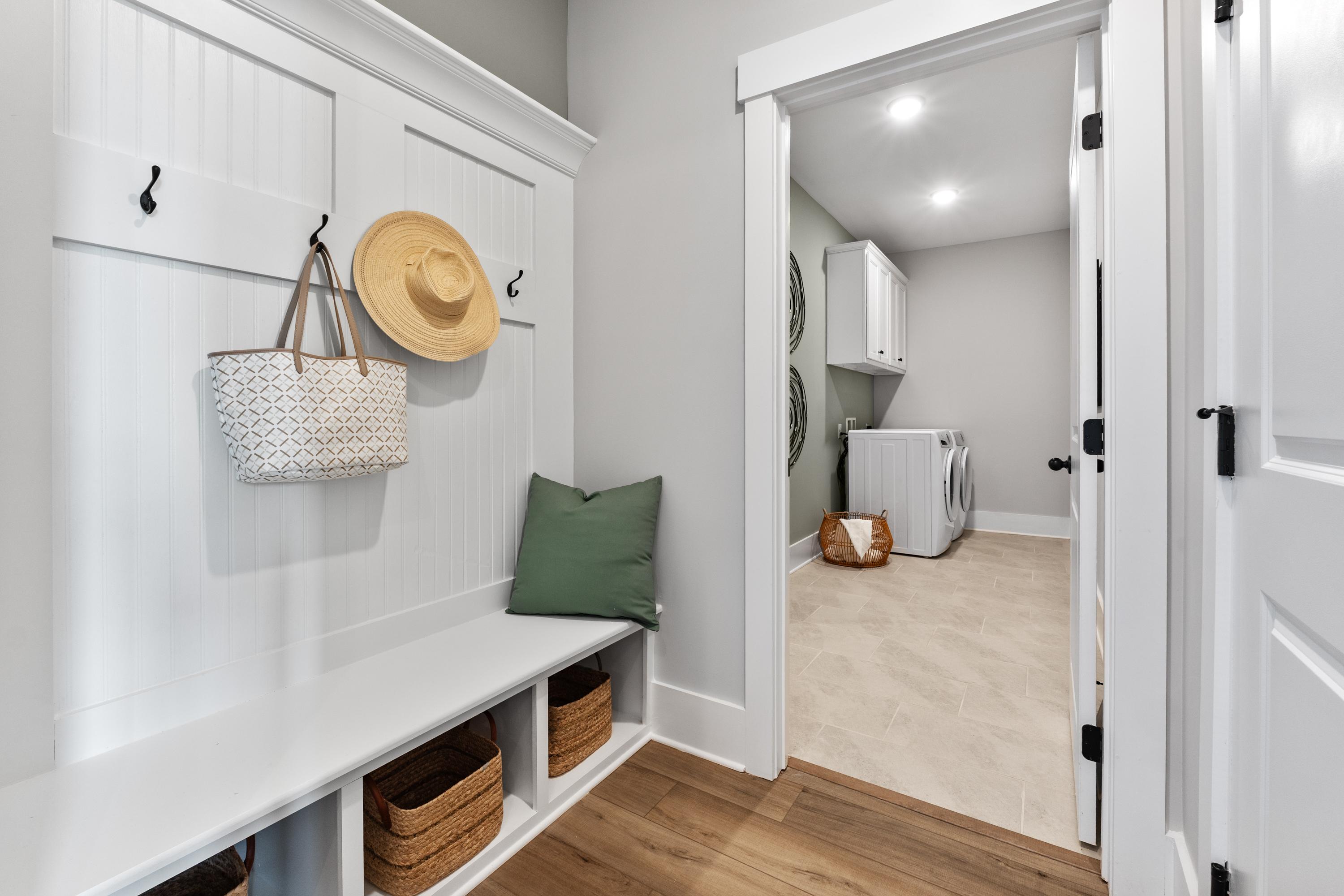 Spacious mudroom with white shiplap hooks, yellow hat, tote bag, built-in bench, wicker baskets, open to laundry room in River Road Estates Decatur AL