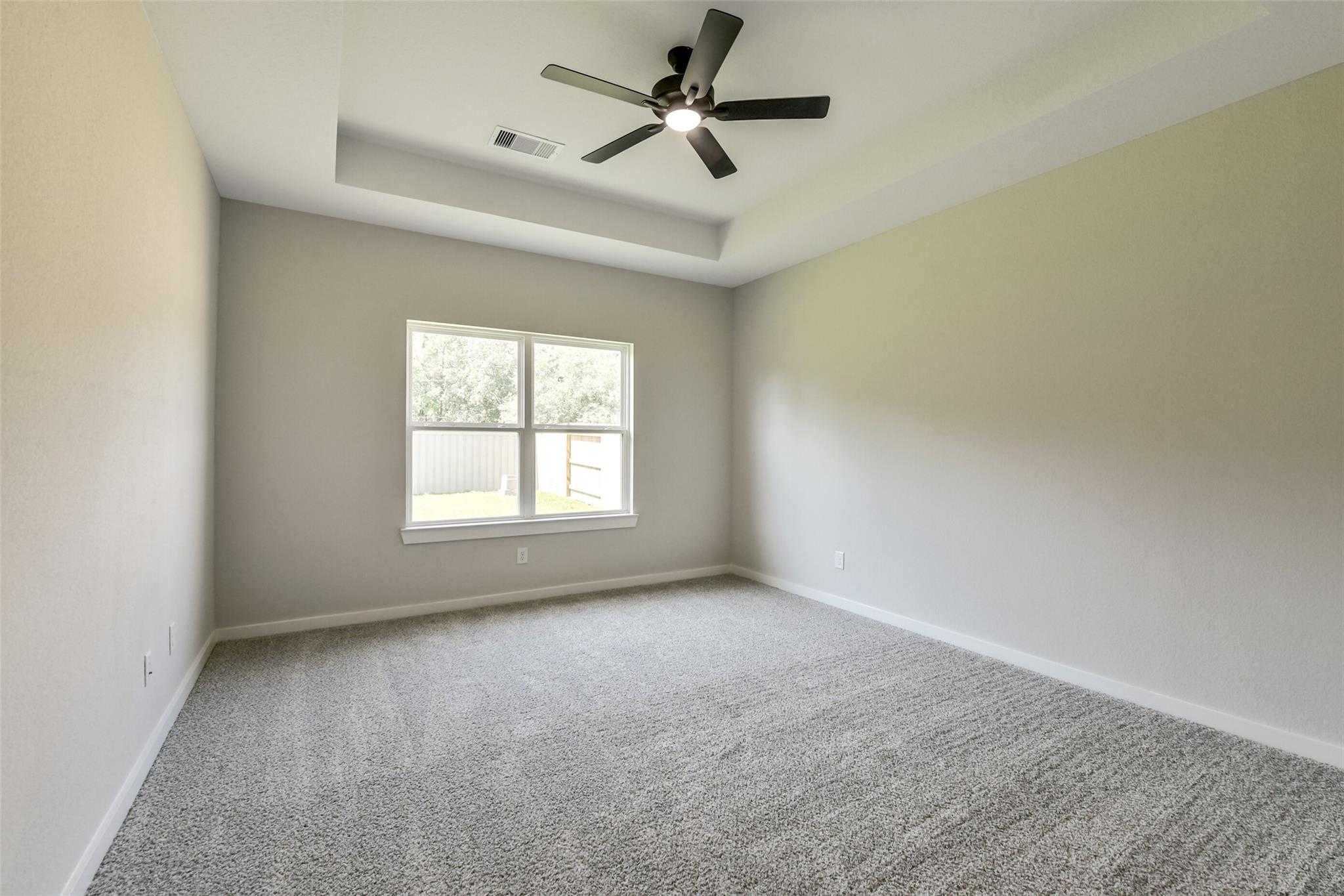 Spacious bedroom with tray ceiling, dual fans, beige walls, and large windows in Davidson Homes The Daphne H, Crosby, Texas