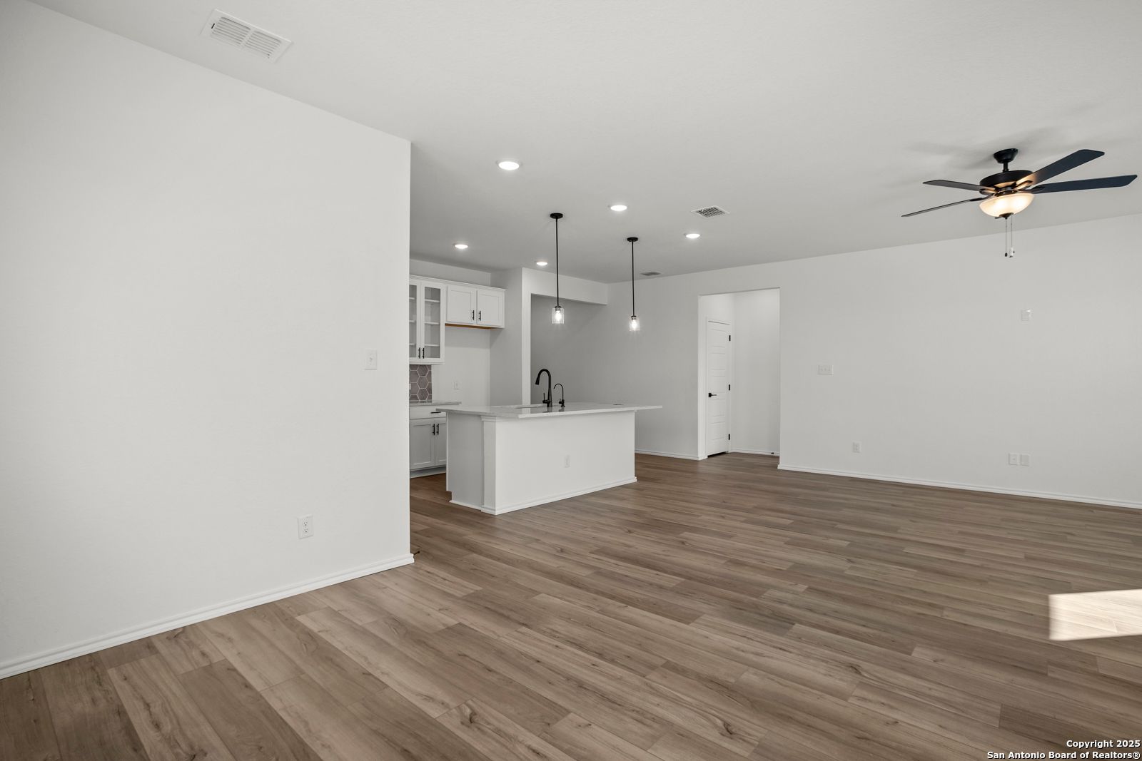 Modern open-concept kitchen with white island sink and cabinets, adjacent living area with hardwood floors and ceiling fan in The Daphne K, San Antonio