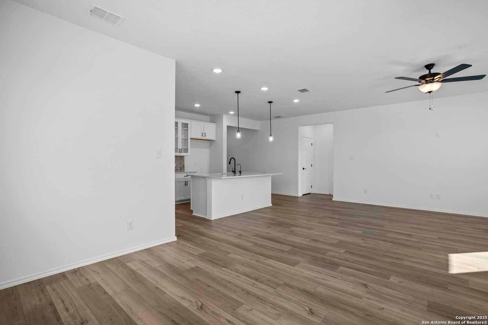 Modern open-concept kitchen with white island sink and cabinets, adjacent living area with hardwood floors and ceiling fan in The Daphne K, San Antonio