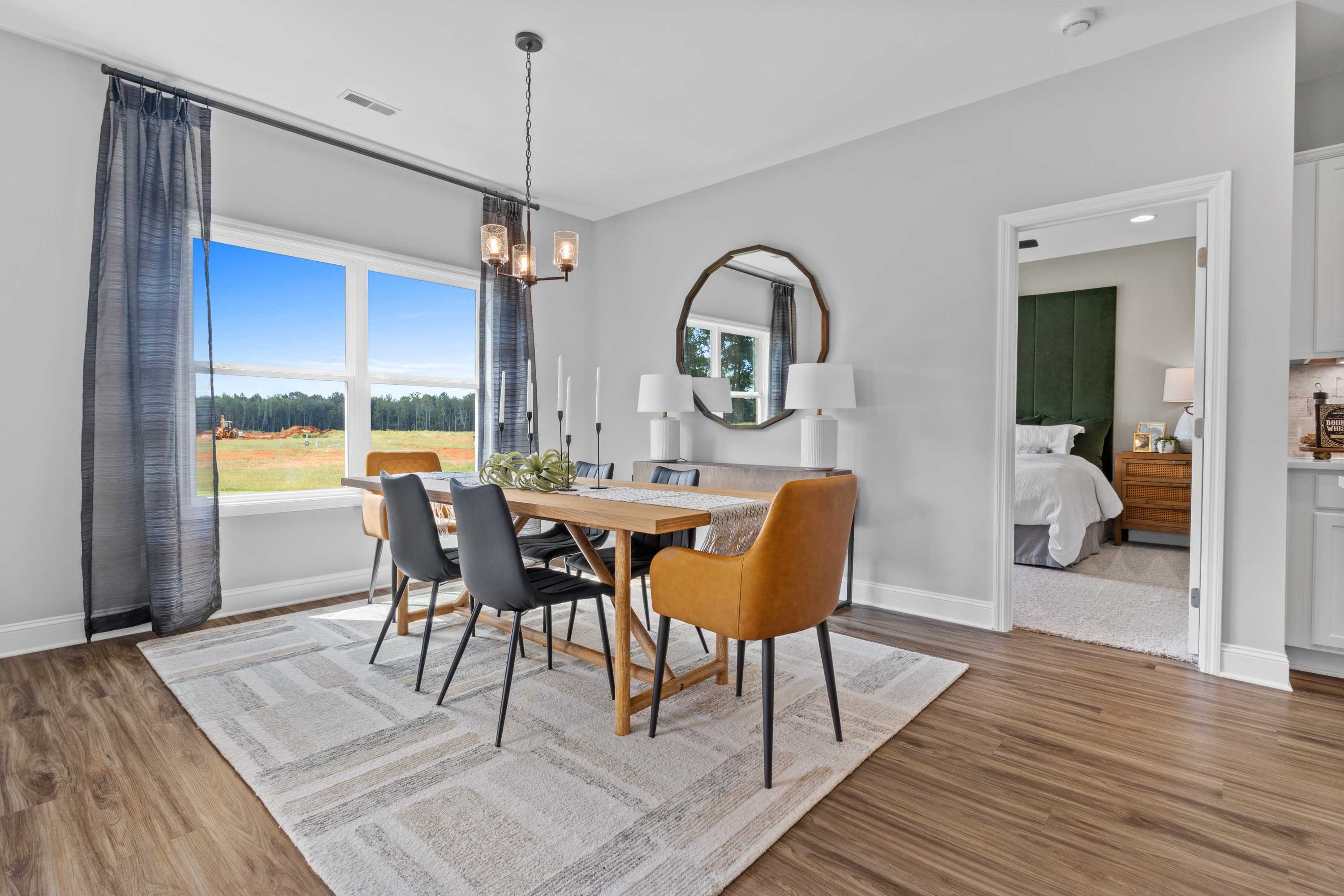 Spacious dining room in Wood Trail Toney Alabama with wooden table, orange chairs, round mirror, chandelier, and field views through large windows