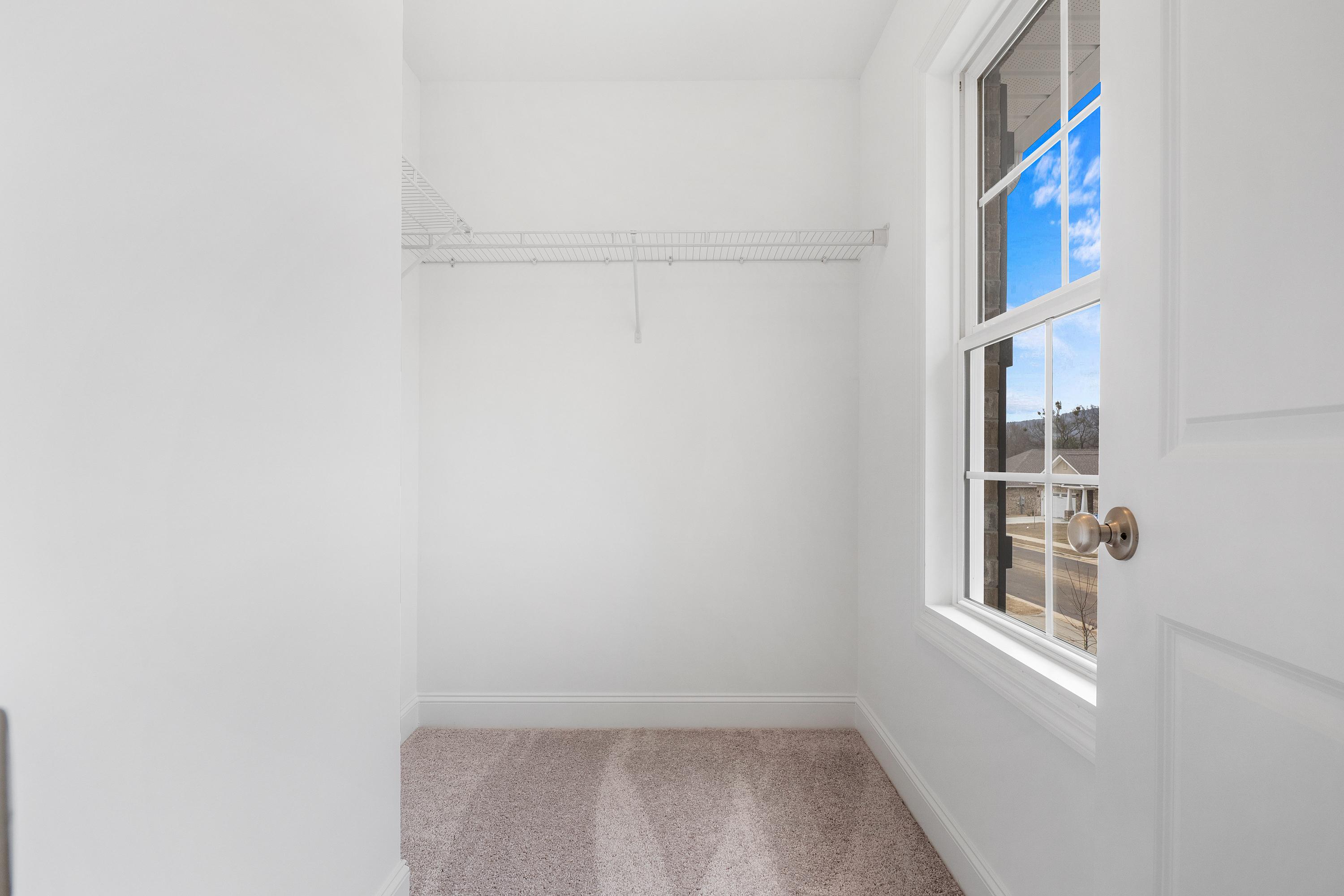 Spacious walk-in master closet in The Aiken by Davidson Homes with white walls, hanging rod, beige carpet, and window overlooking blue sky