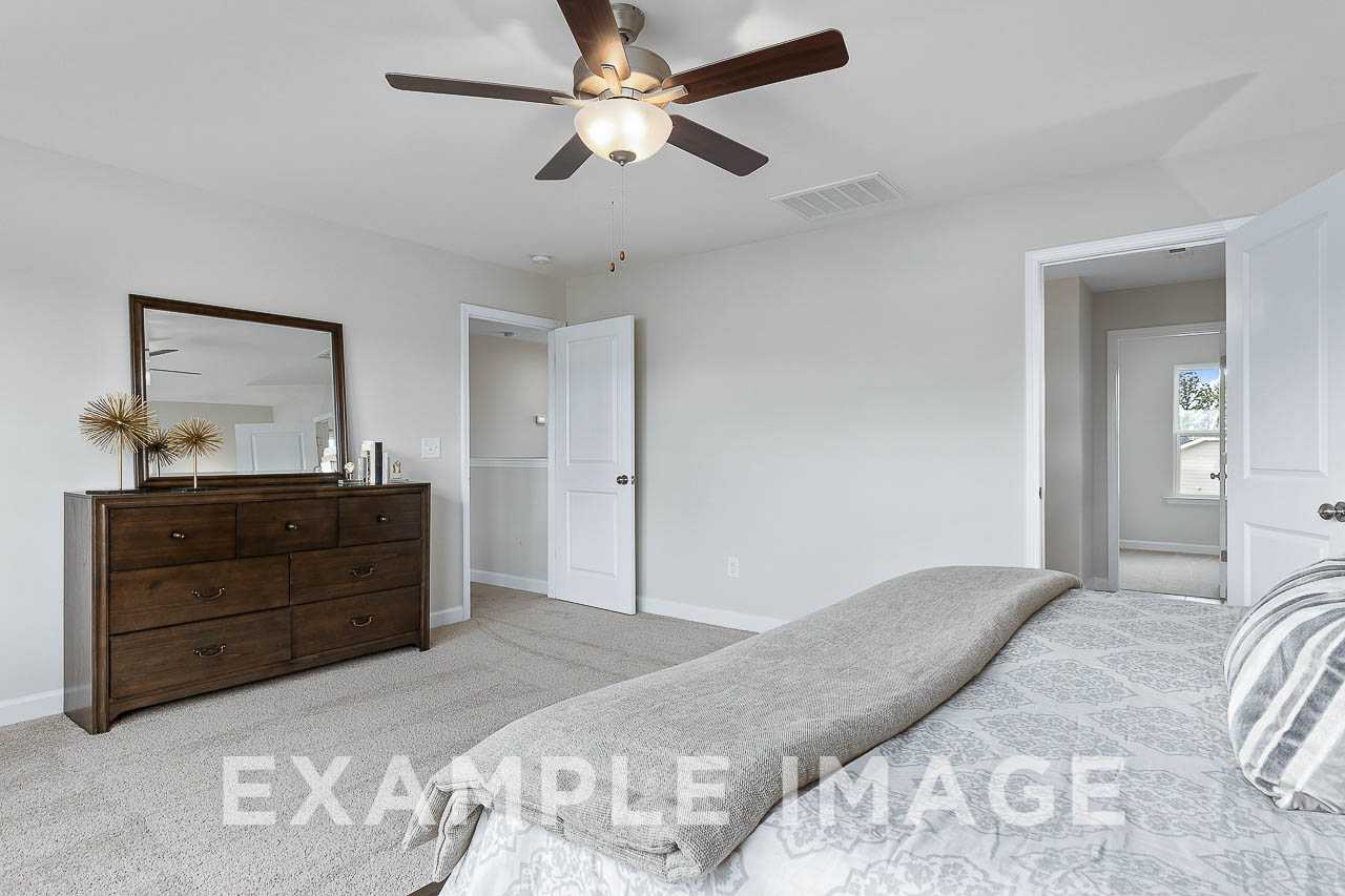 Spacious master bedroom in The Willow C featuring wooden dresser, king bed with gray linens, ceiling fan, and en-suite bath door