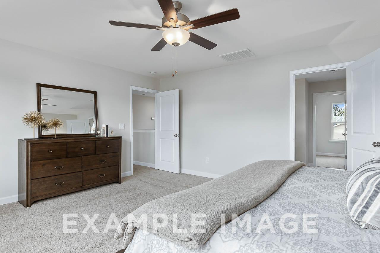 Spacious master bedroom in The Willow C featuring wooden dresser, king bed with gray linens, ceiling fan, and en-suite bath door