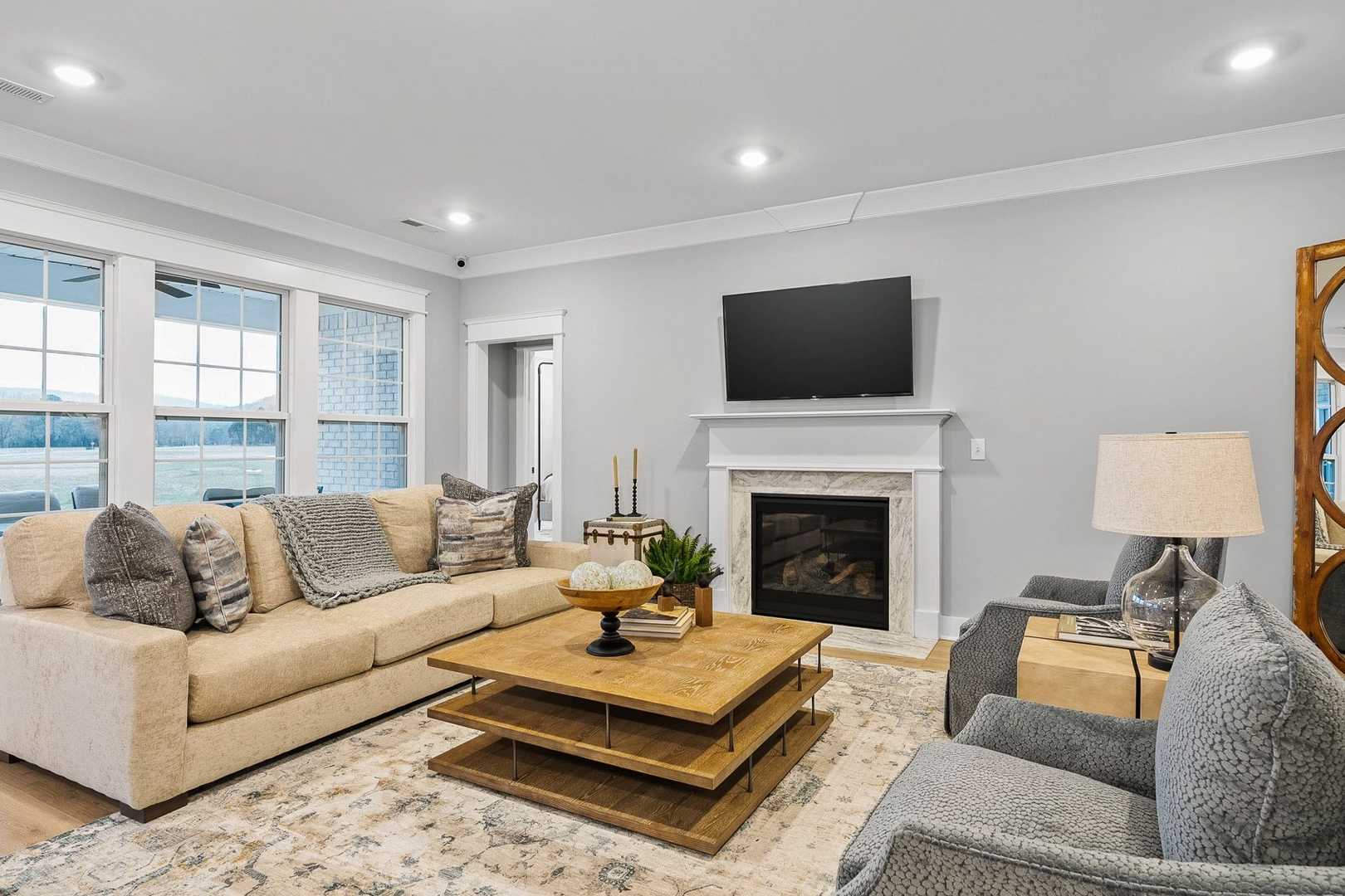 Cozy living room in The Oxford home with beige L-shaped sofa, wooden coffee table, gas fireplace, mounted TV, and large windows