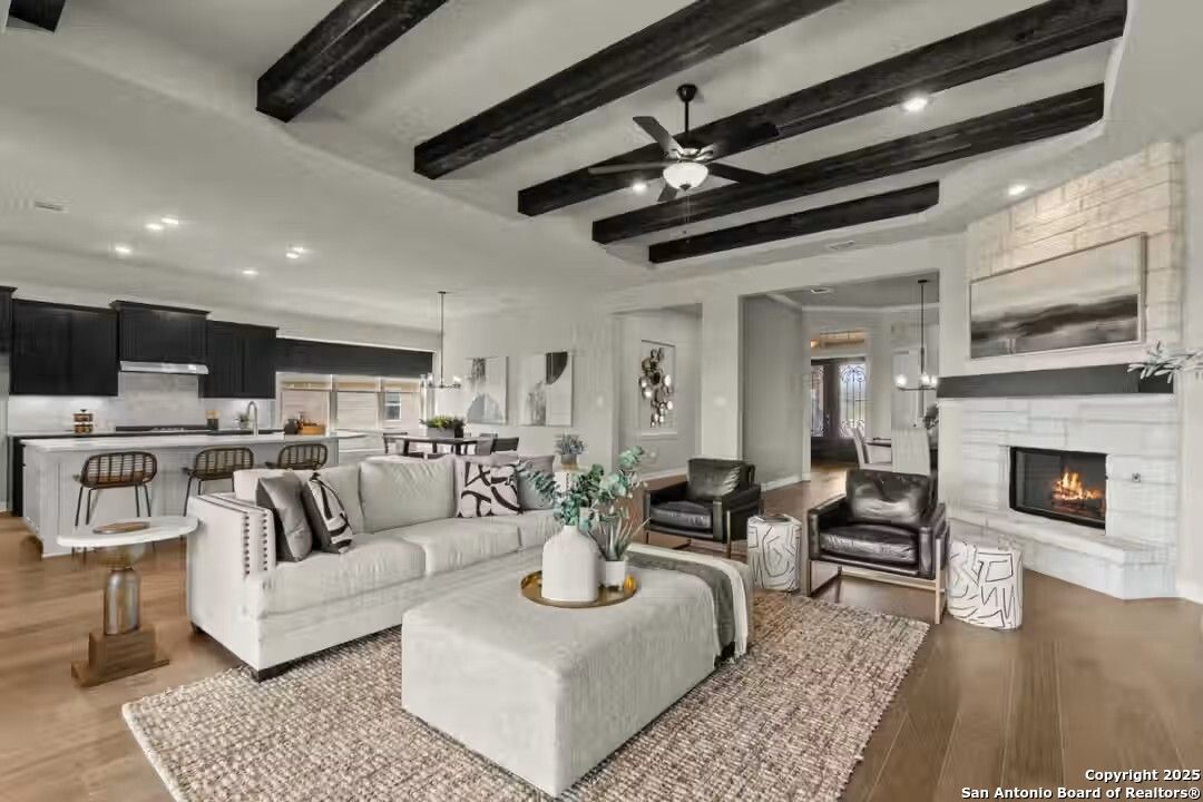 Modern open-concept living room with stone gas fireplace, exposed beam ceiling, white sofa, and kitchen island in The Summerlin B, Castroville, Texas