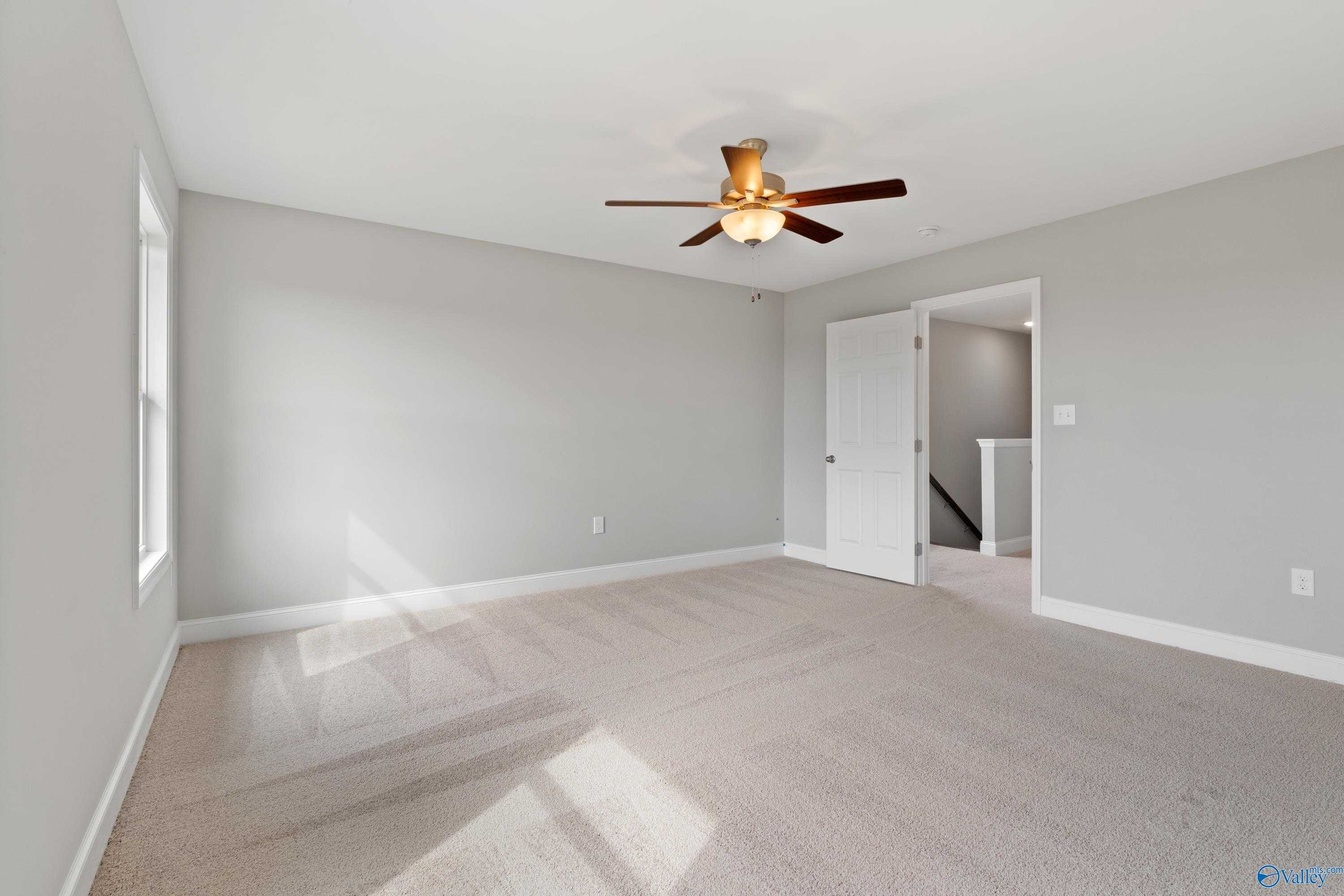 Spacious empty bedroom with gray walls, beige carpet, ceiling fan, and staircase access in The Aiken 3-bedroom home, Meridianville, Alabama